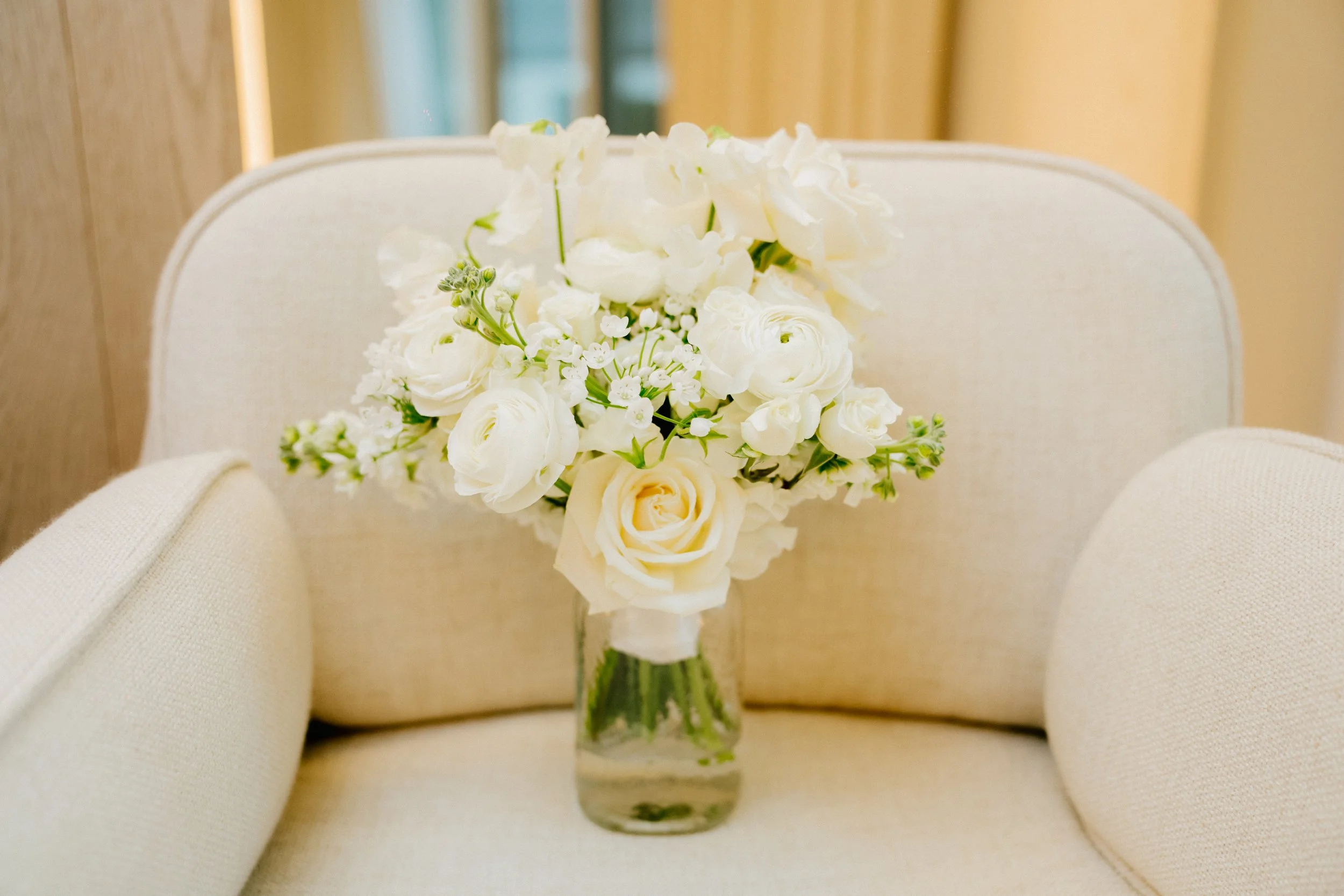 A bouquet of white flowers, including roses and other blooms, is arranged in a small glass vase and placed on a beige upholstered chair.