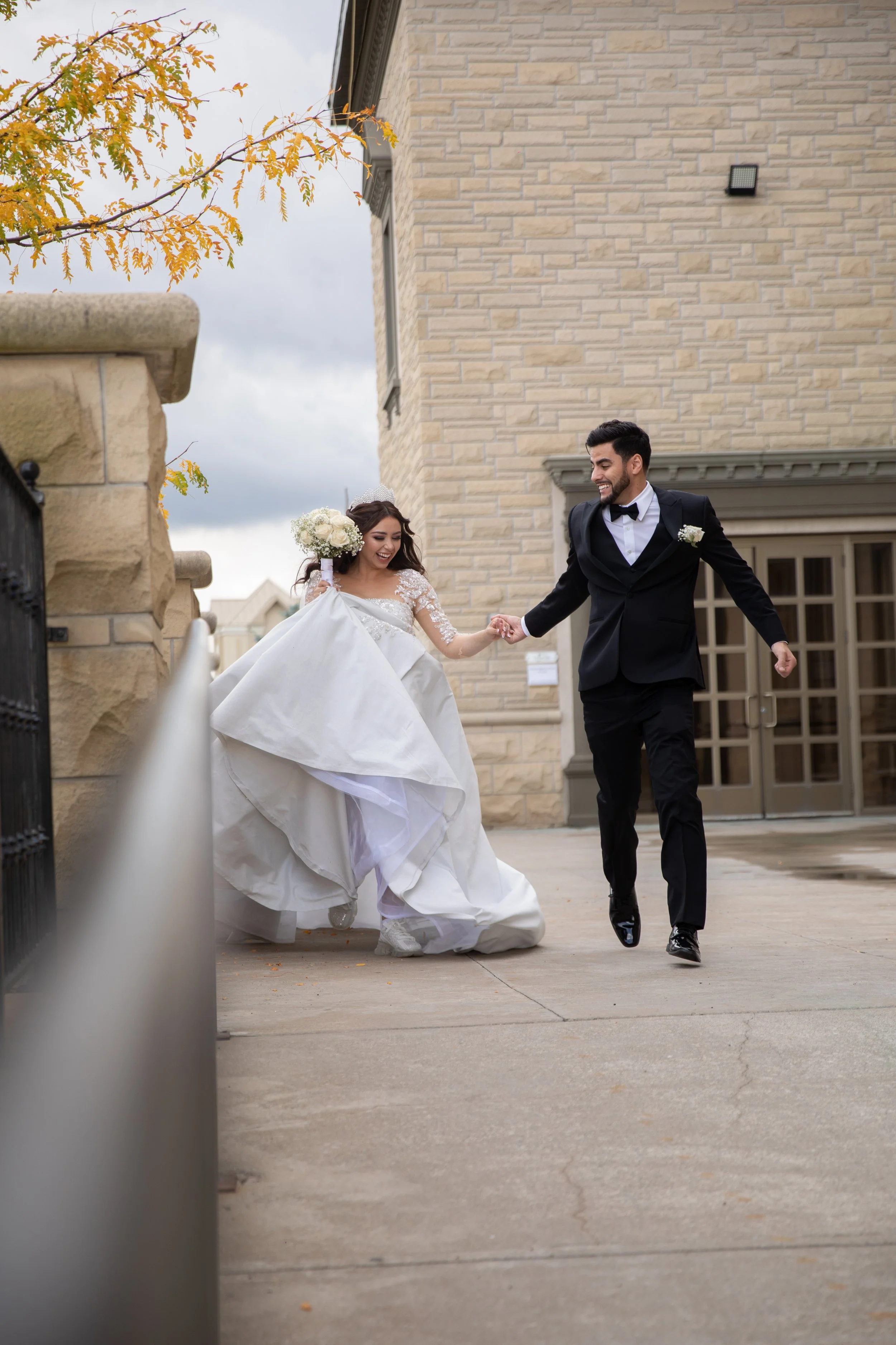 A bride and groom holding hands and smiling outside a building, dressed in wedding attire, with the bride in a white gown and the groom in a black tuxedo.