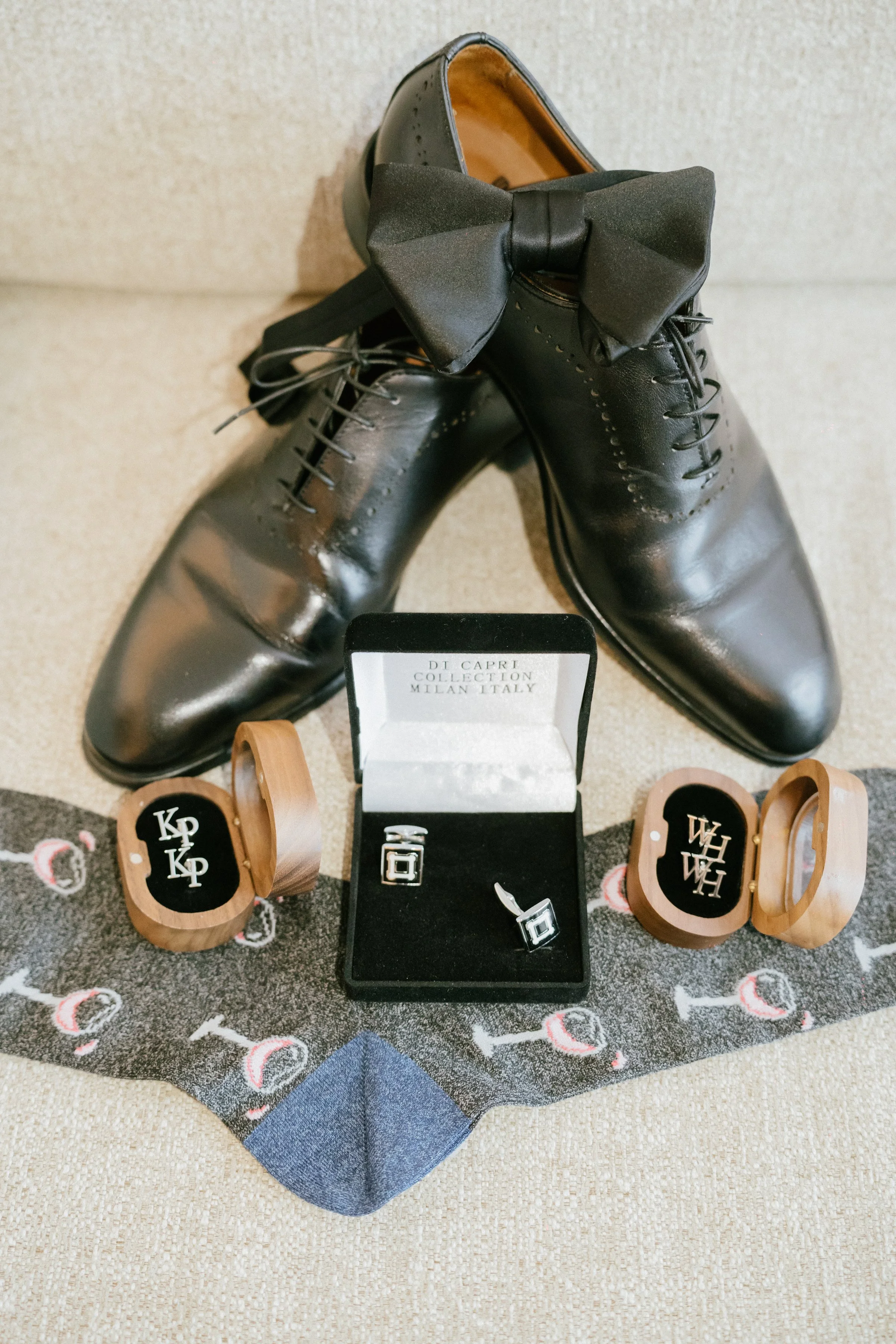 Pair of black leather dress shoes with a black bow tie, a box of silver cufflinks, and two wooden ring boxes with monogrammed rings and wooden cufflinks, all arranged on a patterned scarf.
