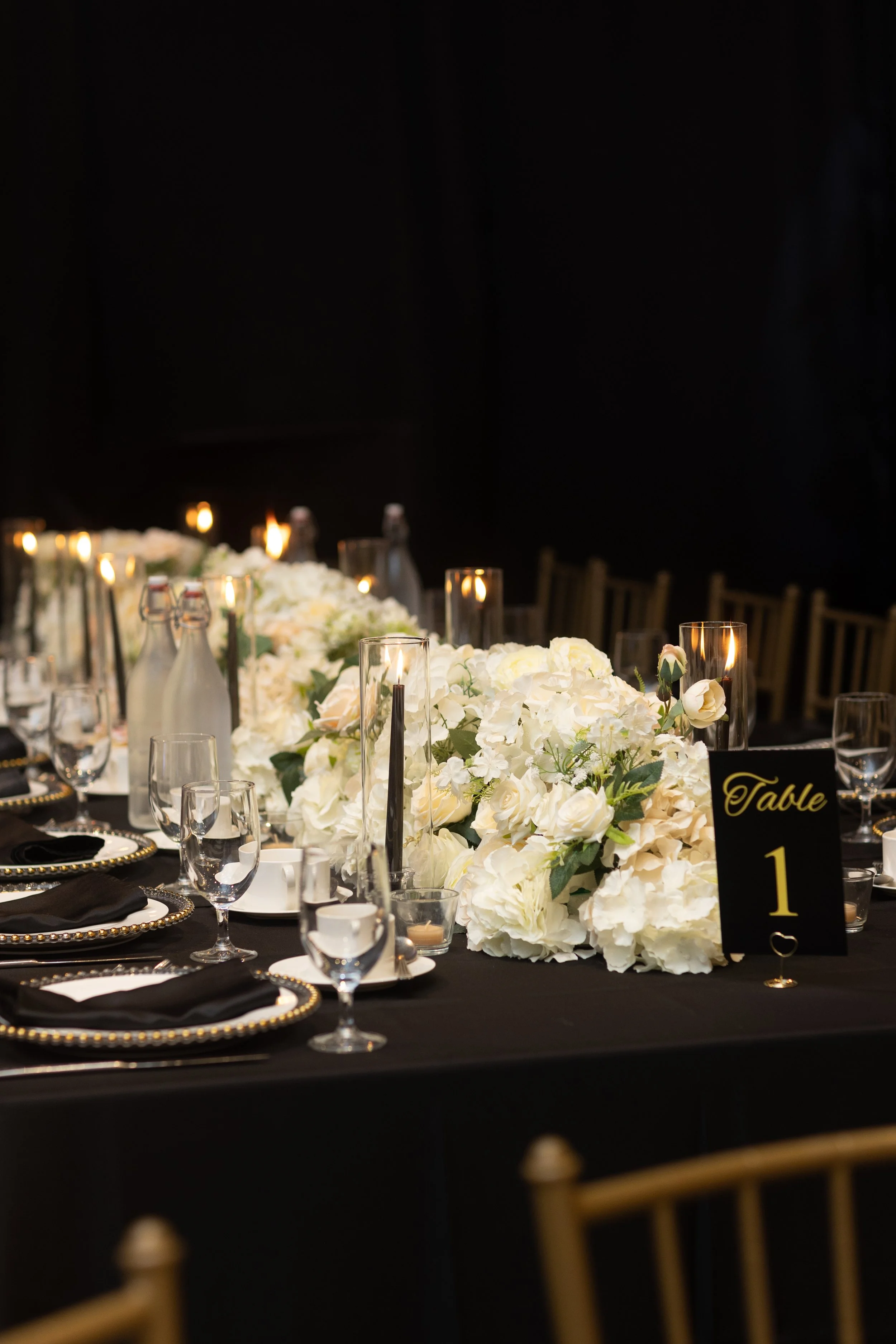 Elegant banquet table decorated with white flowers, candles, black and gold table number sign, and black napkins in a dimly lit room.
