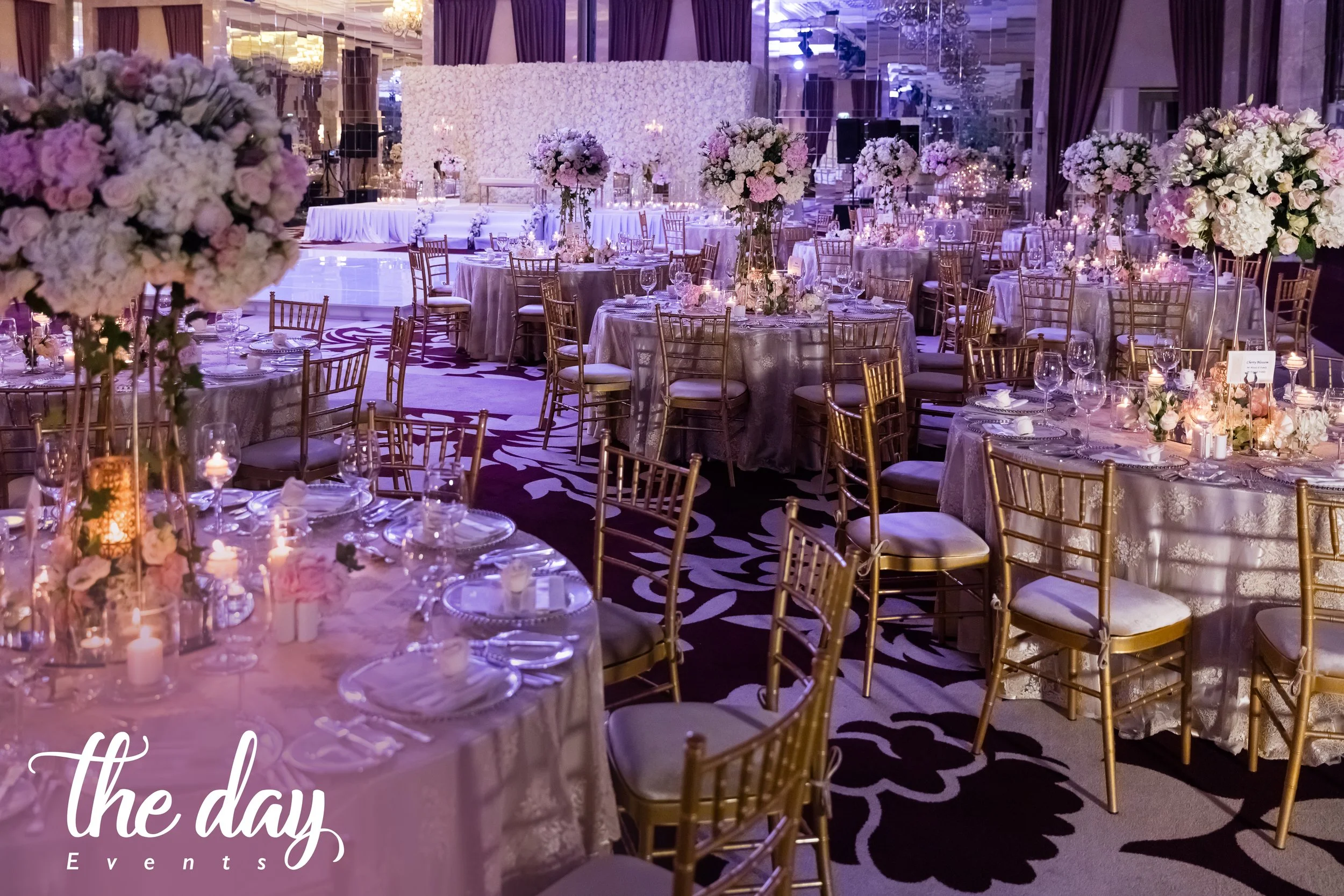 Elegant event reception decorated with gold chairs, round tables with silver tablecloths, and tall floral centerpieces with mostly white and pink flowers. There are candles on the tables, and a floral wall backdrop in the background. The room has pur