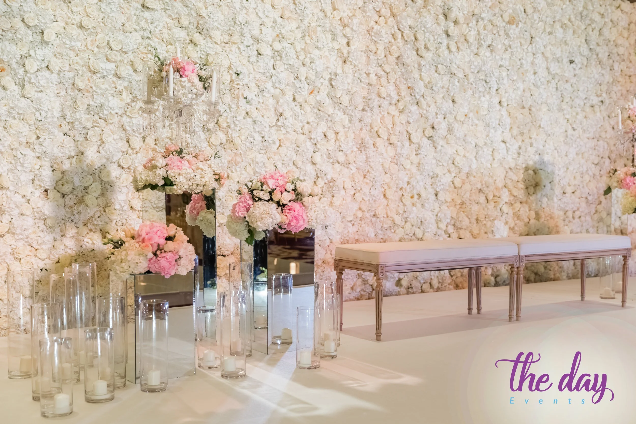 Elegant wedding or event decor with a wall of white and pink flowers, tall mirrored vases with pink and white flowers, glass cylindrical candle holders with white candles, and a light-colored bench against the floral wall. The logo 'the day Events' i