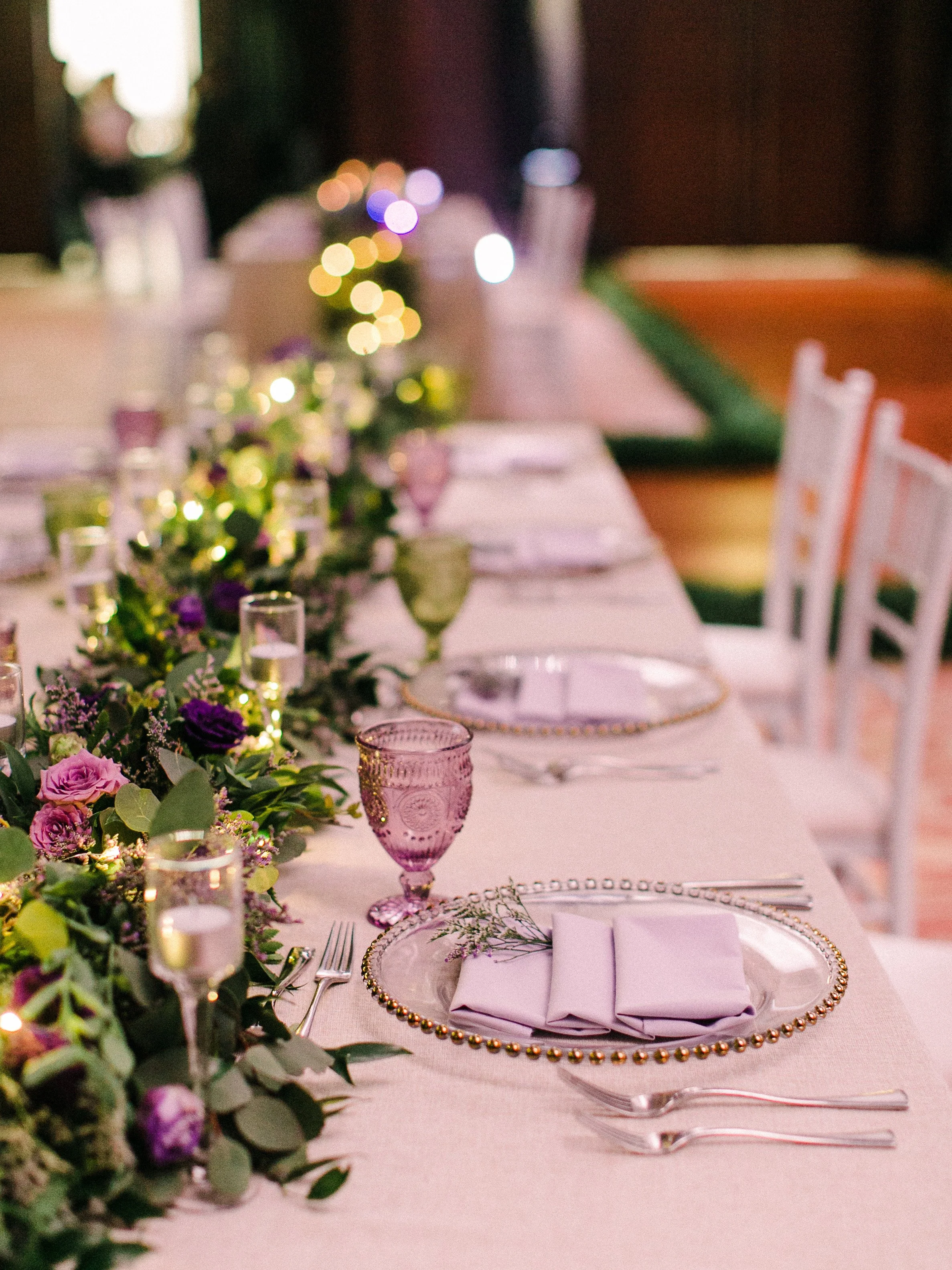 A decorated dining table with purple and pink flowers, purple glassware, white napkins, and gold accents, set for a formal event or celebration.