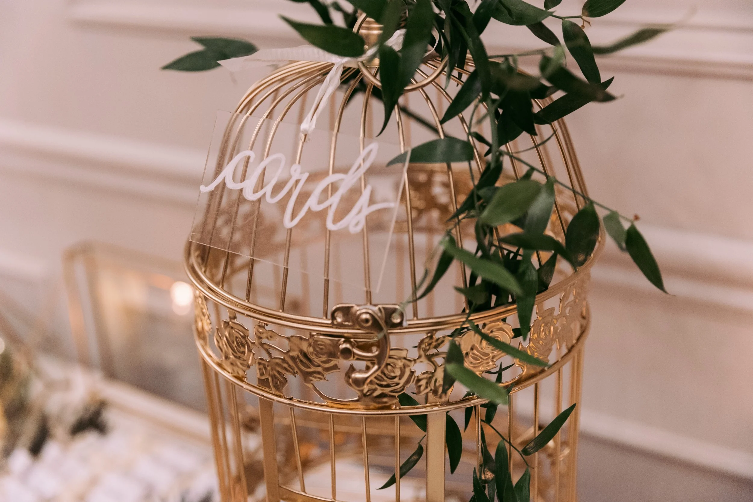 A gold decorative birdcage with a greenery plant inside and a transparent tag that says 'cards' hanging on it.