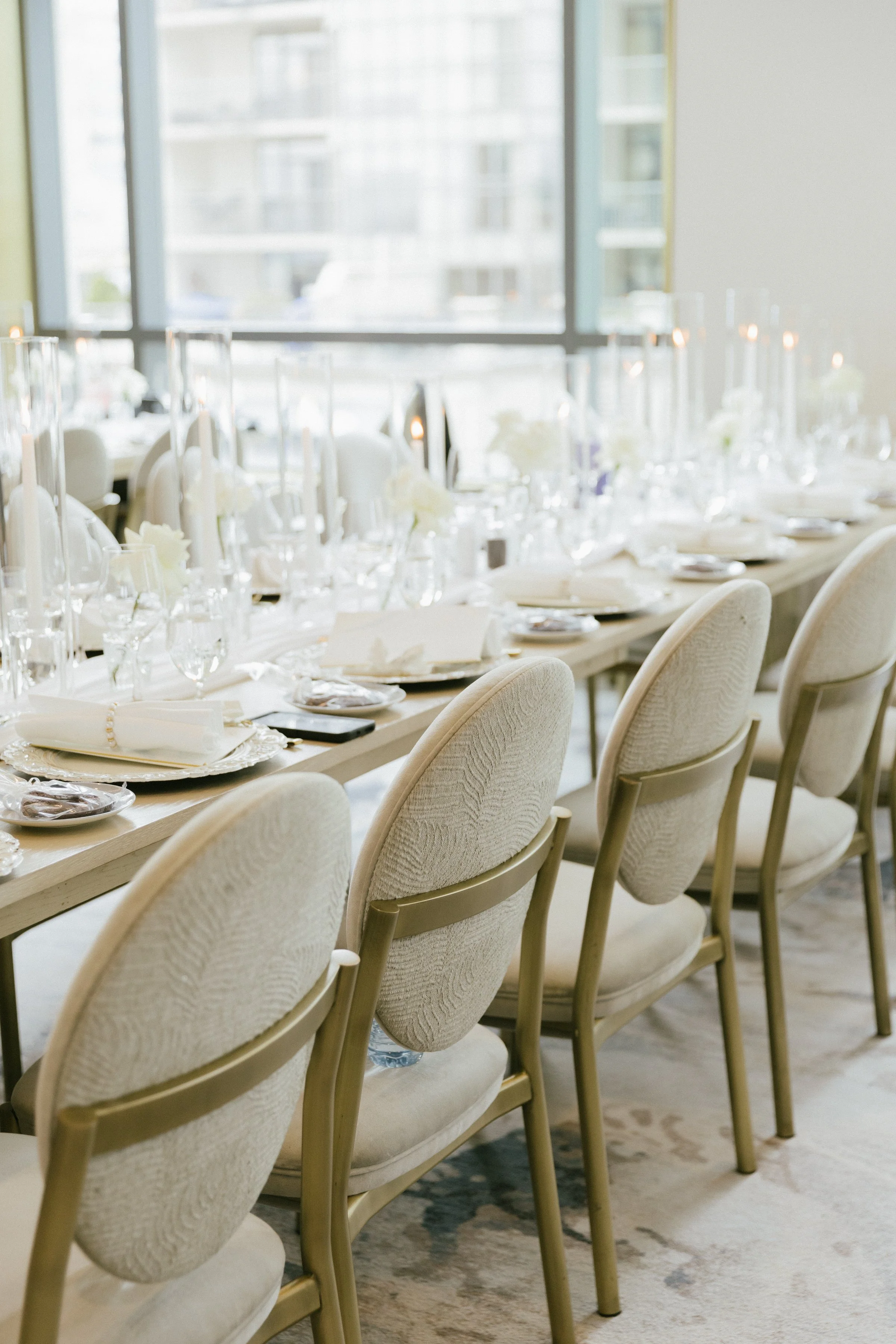 Elegant banquet table set in a bright, modern room with large windows, featuring white chairs with gold frames, white tablecloth, floral centerpieces, candles, and tableware.