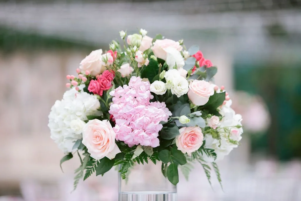 A floral arrangement with pink, white, and cream roses, hydrangeas, and greenery in a glass vase.