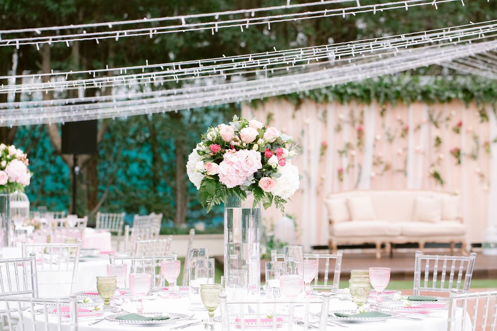 Elegant outdoor event setup with white tables, pink and white floral centerpieces, and white chairs under string lights.
