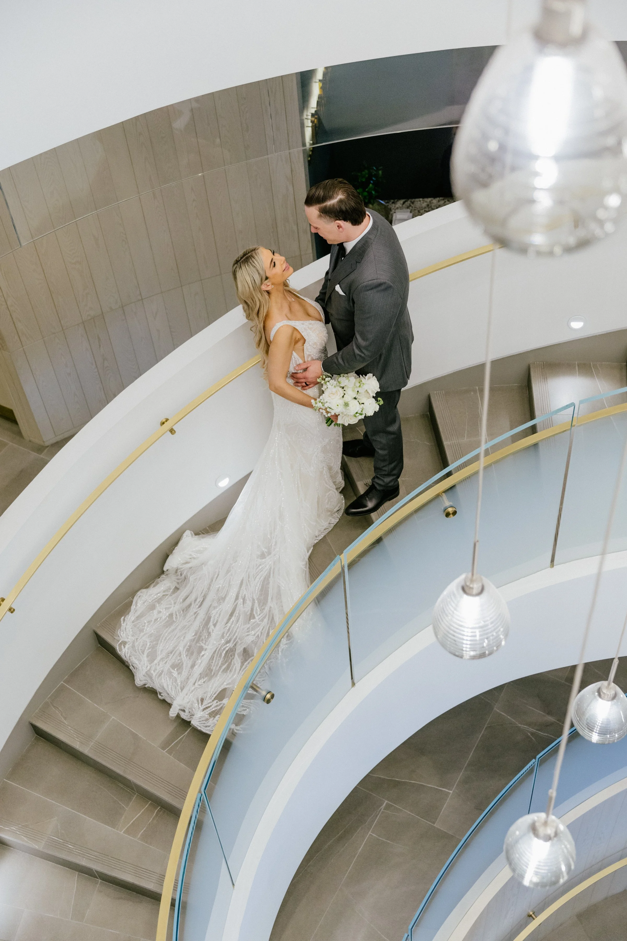 A bride and groom standing on a curved staircase, looking into each other's eyes. The bride is holding a bouquet of white flowers, and the groom has his hands on her waist. The photo is taken from above, with modern lighting fixtures hanging from the