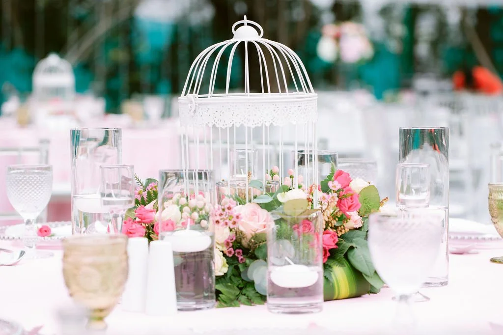 Wedding reception table centerpiece with a white birdcage surrounded by pink and white flowers, candles, and glassware at an outdoor event.