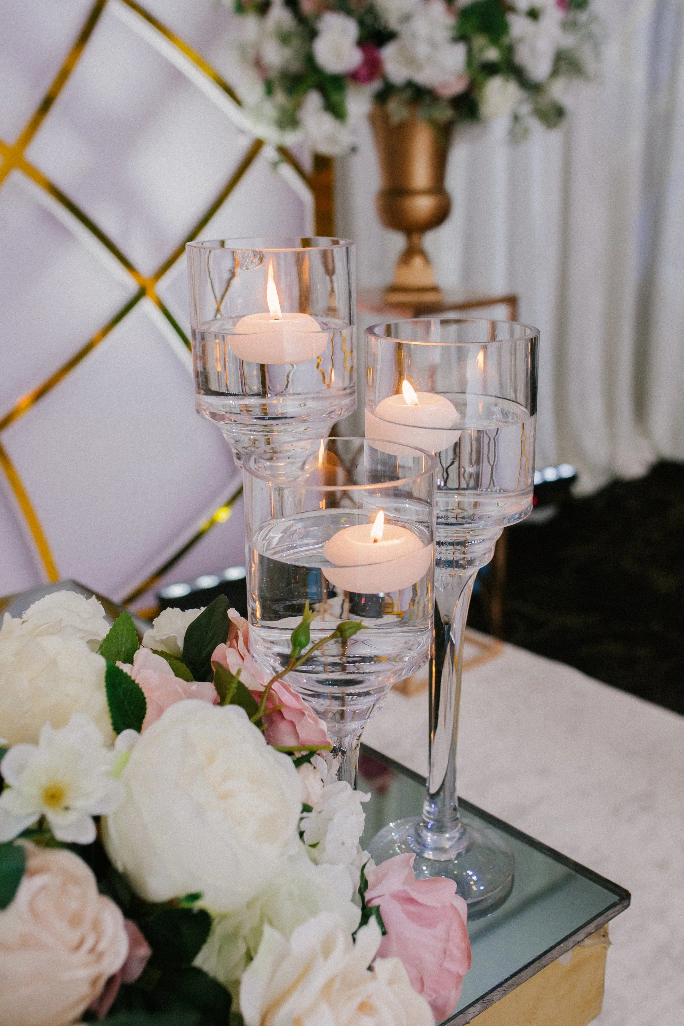 Three tall glass candle holders with floating candles, surrounded by a bouquet of white and pink flowers, on a glass-topped table in an elegant setting.