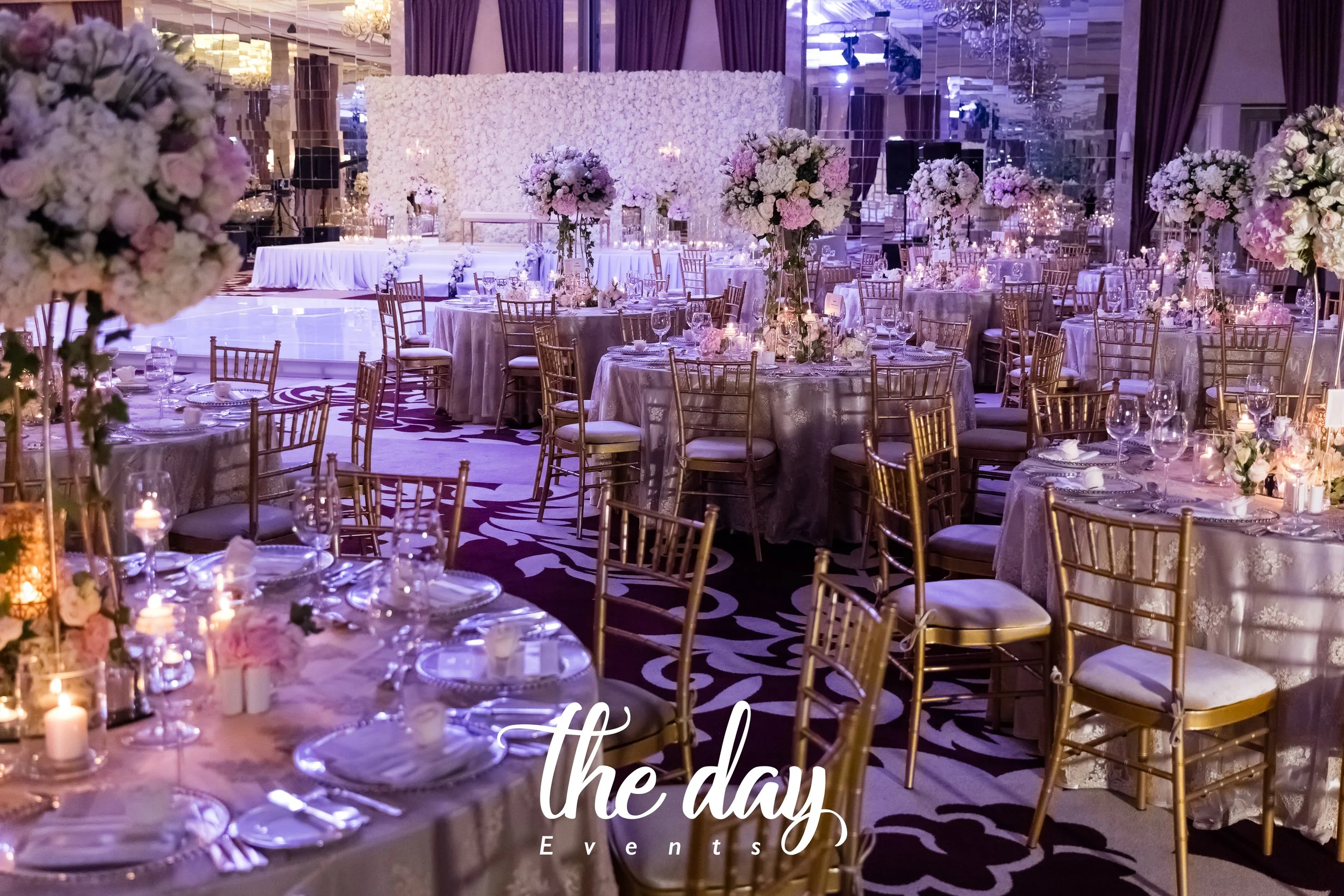 Elegant banquet hall decorated for a wedding or special event, with round tables covered in silver lace tablecloths, tall floral centerpieces with white and pink flowers, gold chairs, and candlelit table settings. The background features a white flor