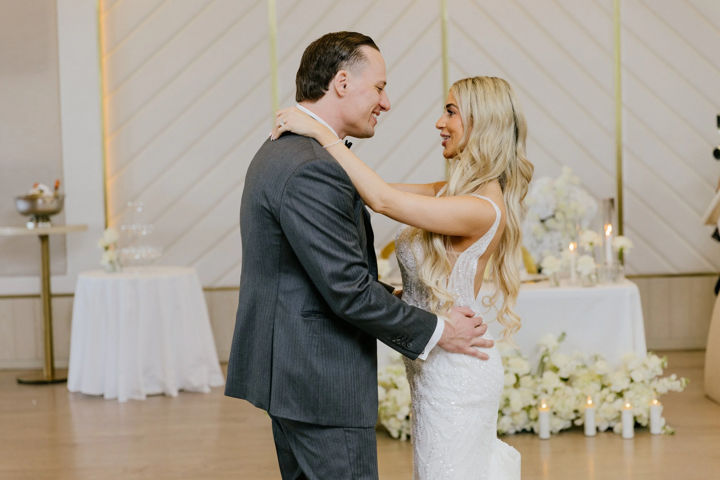 A bride and groom dance during their wedding reception, gazing at each other lovingly in a decorated venue with floral arrangements and candles in the background.