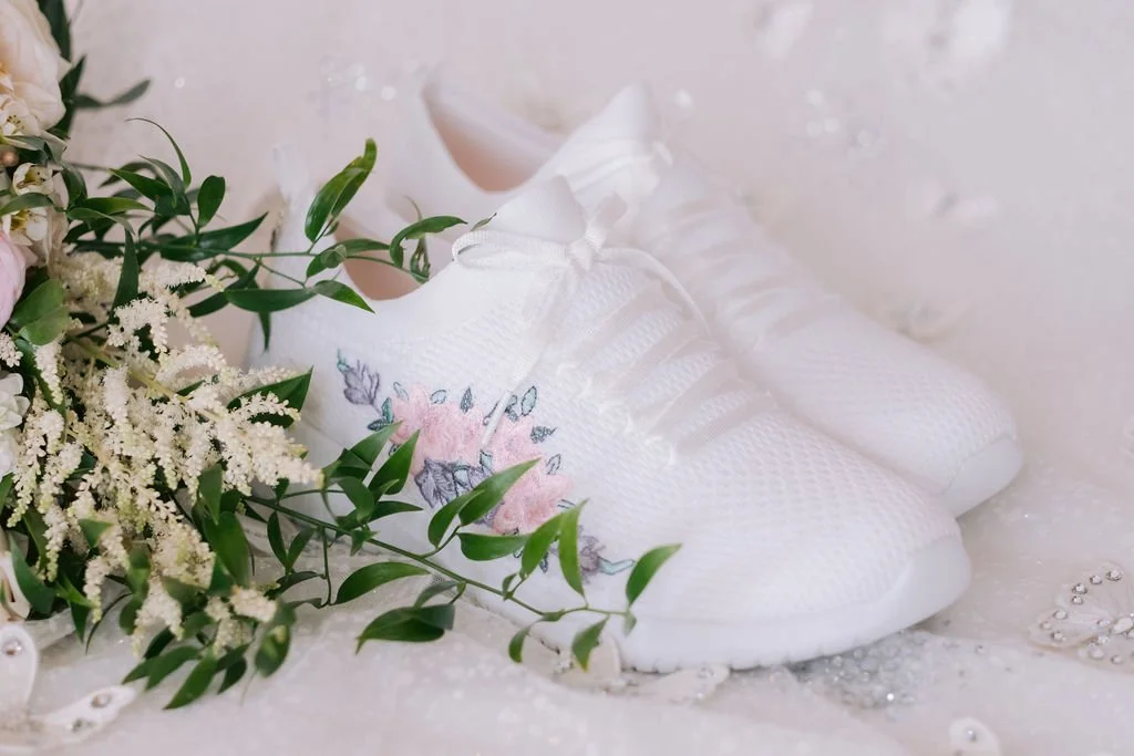 White embroidered sneakers with pink floral design on a white surface next to white flowers and green leaves.