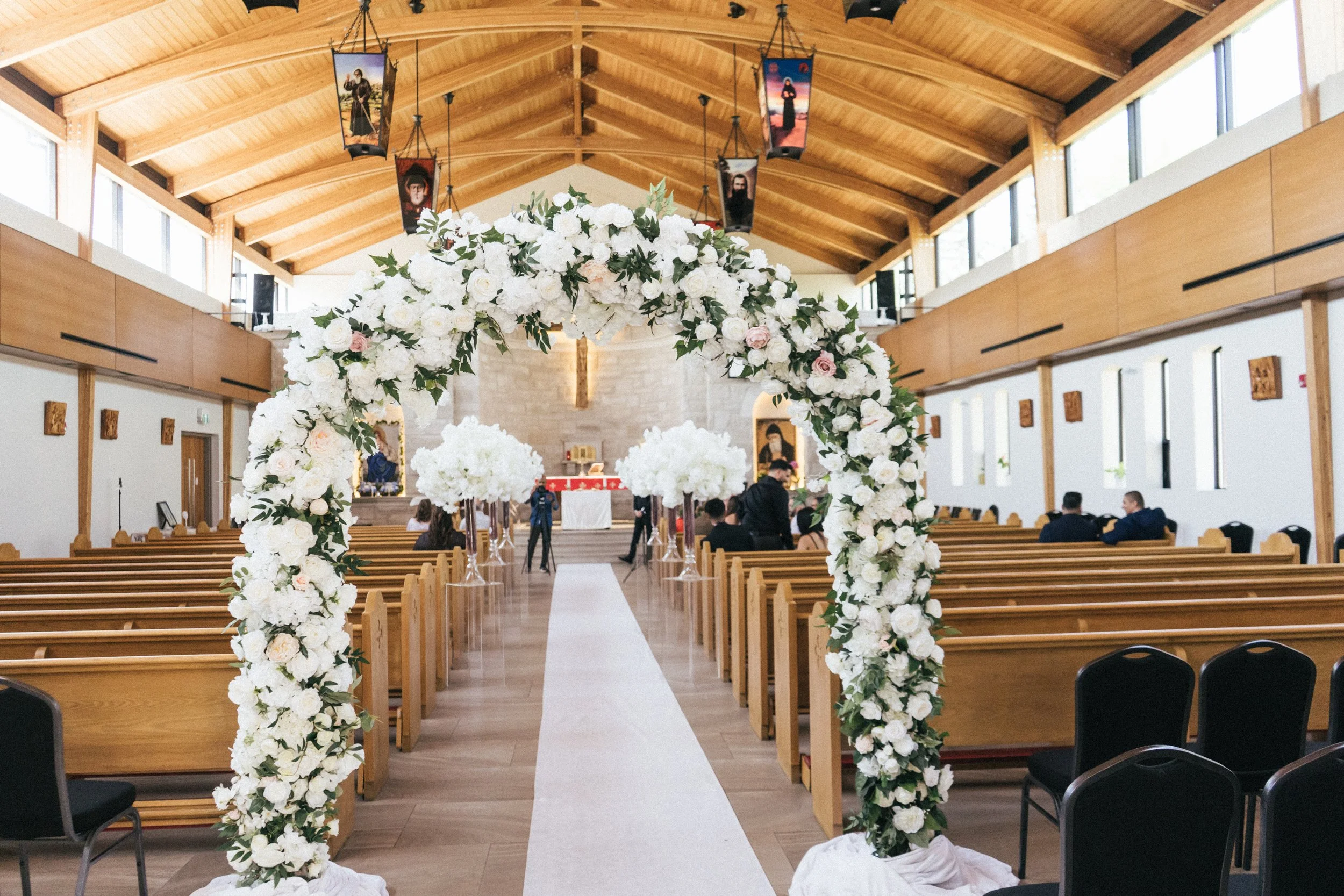 Interior of a church decorated for a wedding, featuring a floral arch of white and light pink flowers, a white aisle runner, and rows of wooden pews.