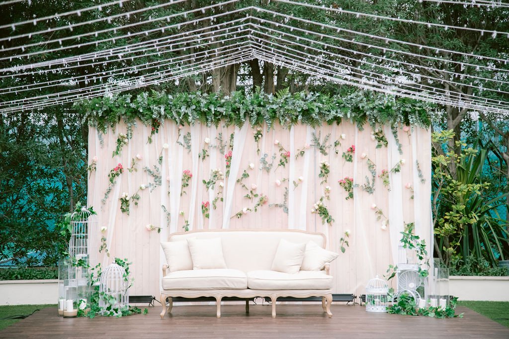 Decorative white wedding or event backdrop with greenery and flowers, a white vintage-style sofa, and birdcages with candles, set outdoors under string lights.