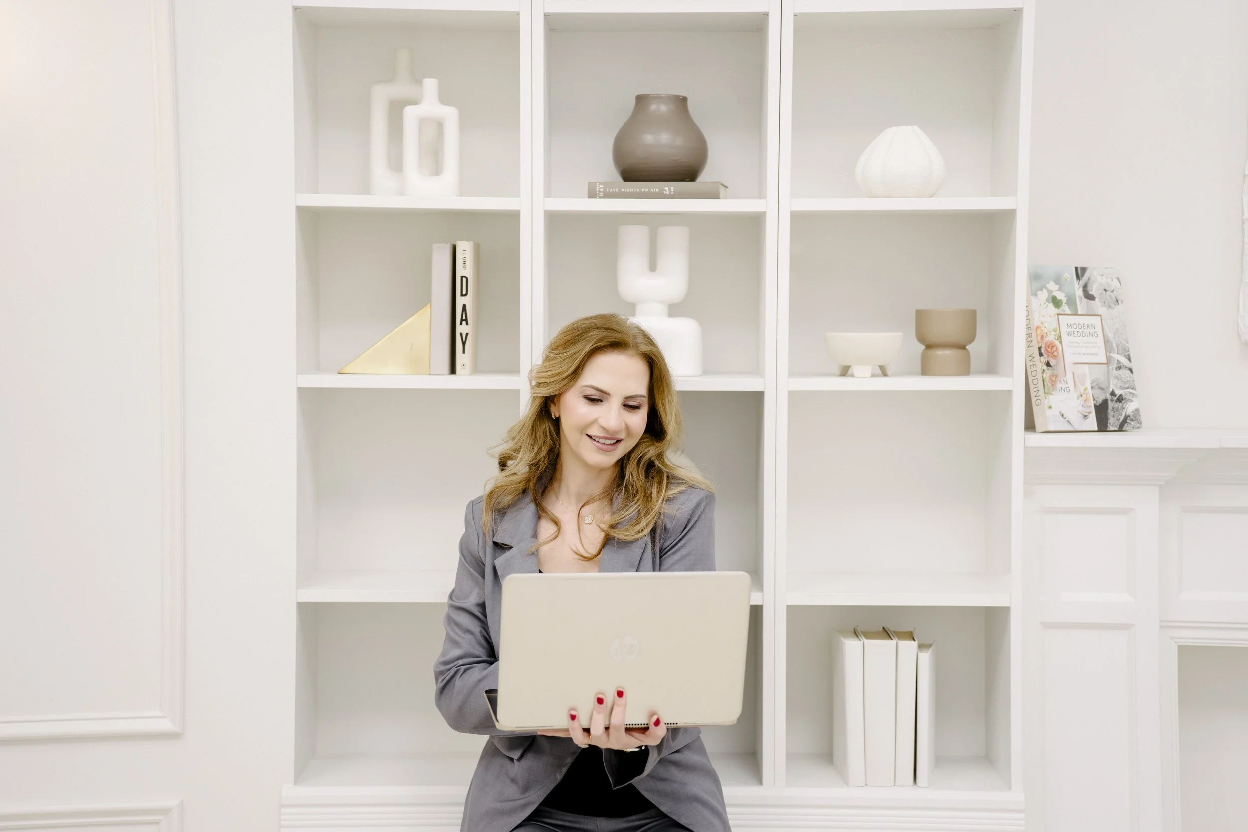 A woman in a gray blazer smiling while looking at a laptop in a white room with decorative shelves and books behind her.