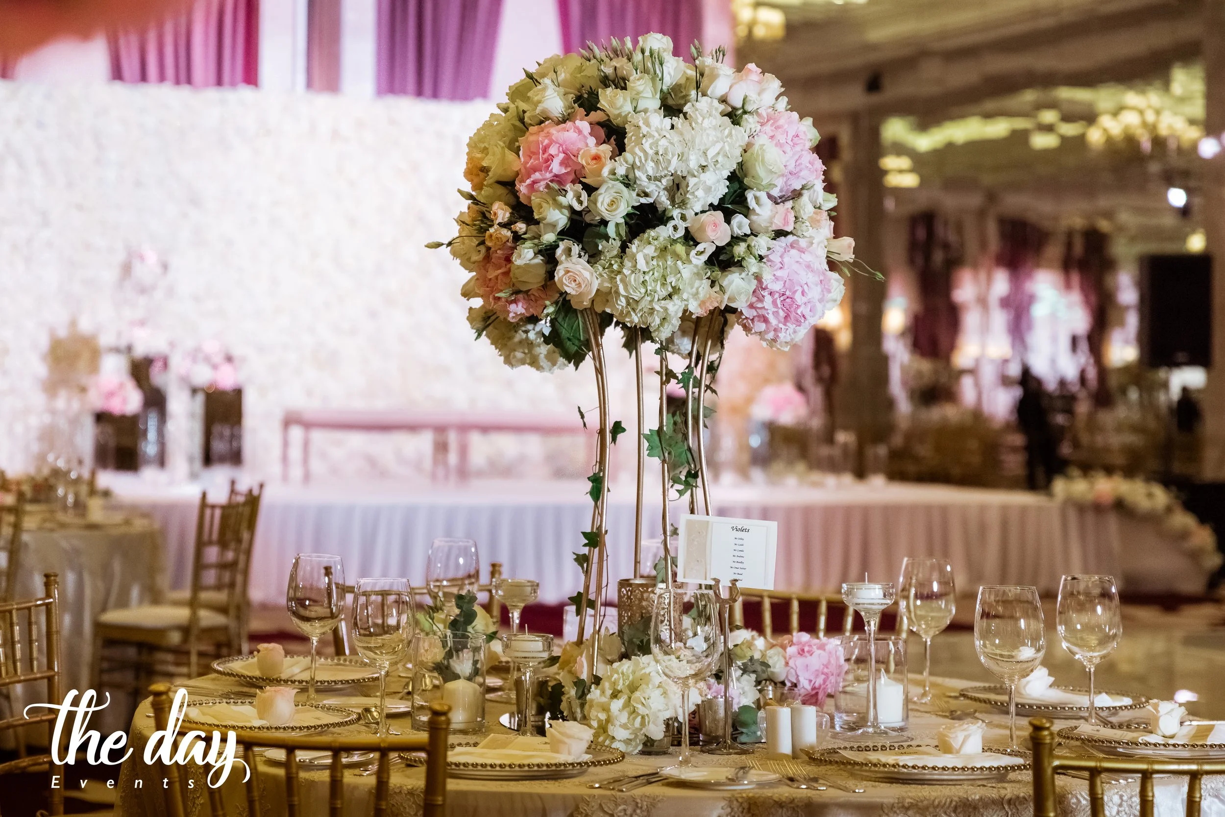 Elegant wedding reception table with a large floral centerpiece of white and pink flowers, surrounded by gold chairs, glassware, and candles. In the background, a decorated reception hall with pink drapes and floral arrangements.