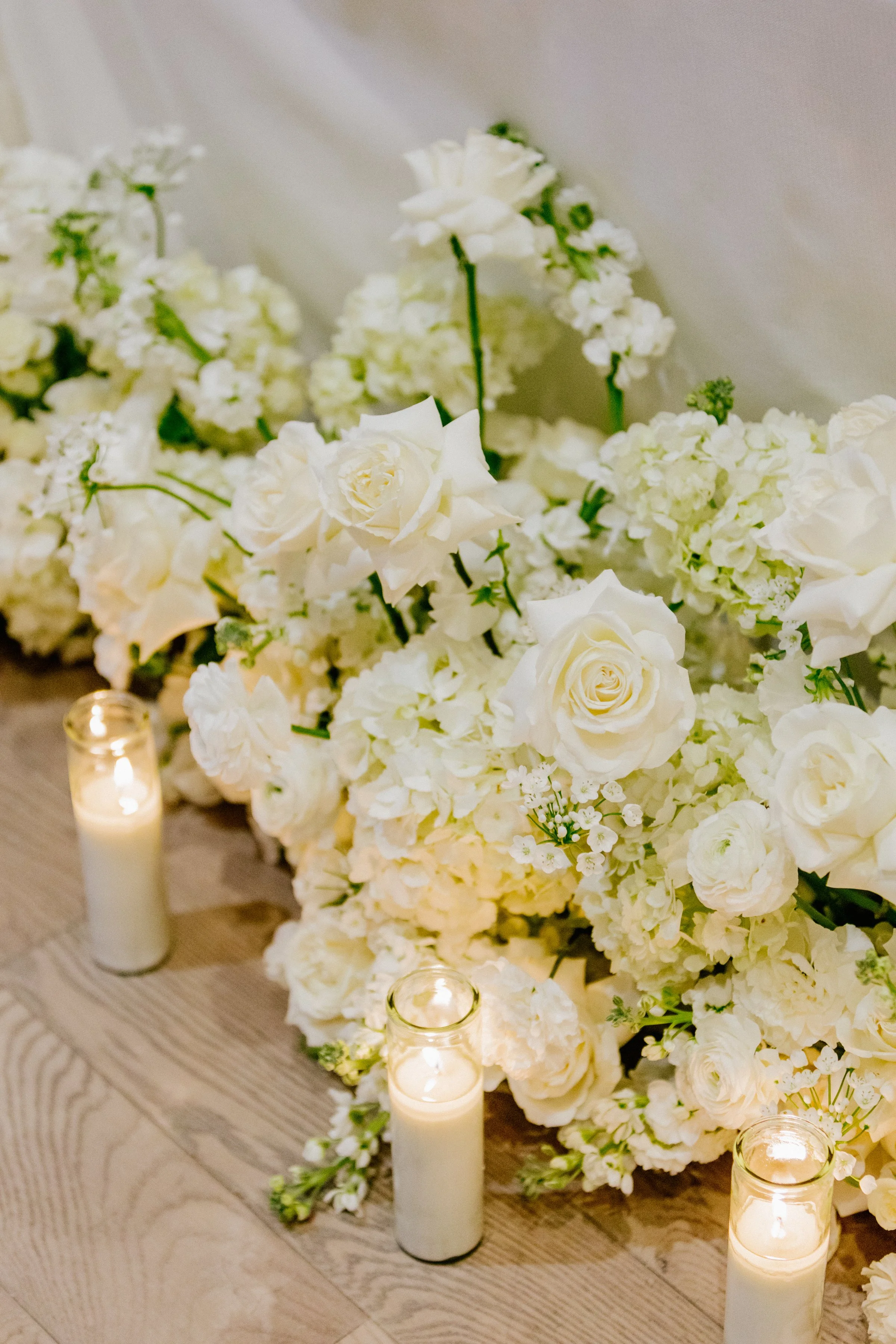 Wedding or event floral arrangement with white roses, hydrangeas, and other white flowers, decorated with lit candles in glass holders on a wooden surface.
