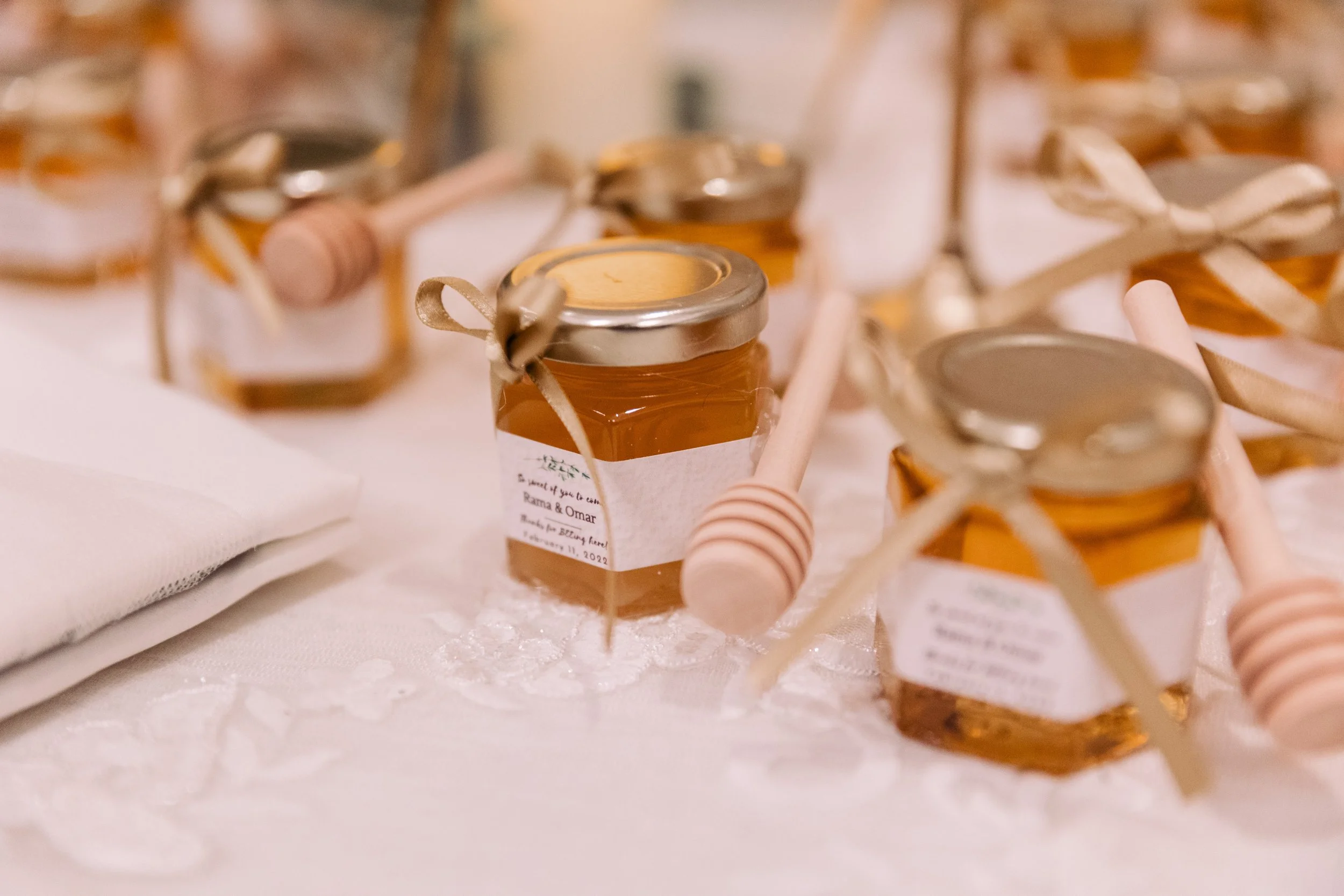 Small jars of honey with pink honey dippers on a white cloth, some jars have ribbons tied around their lids.