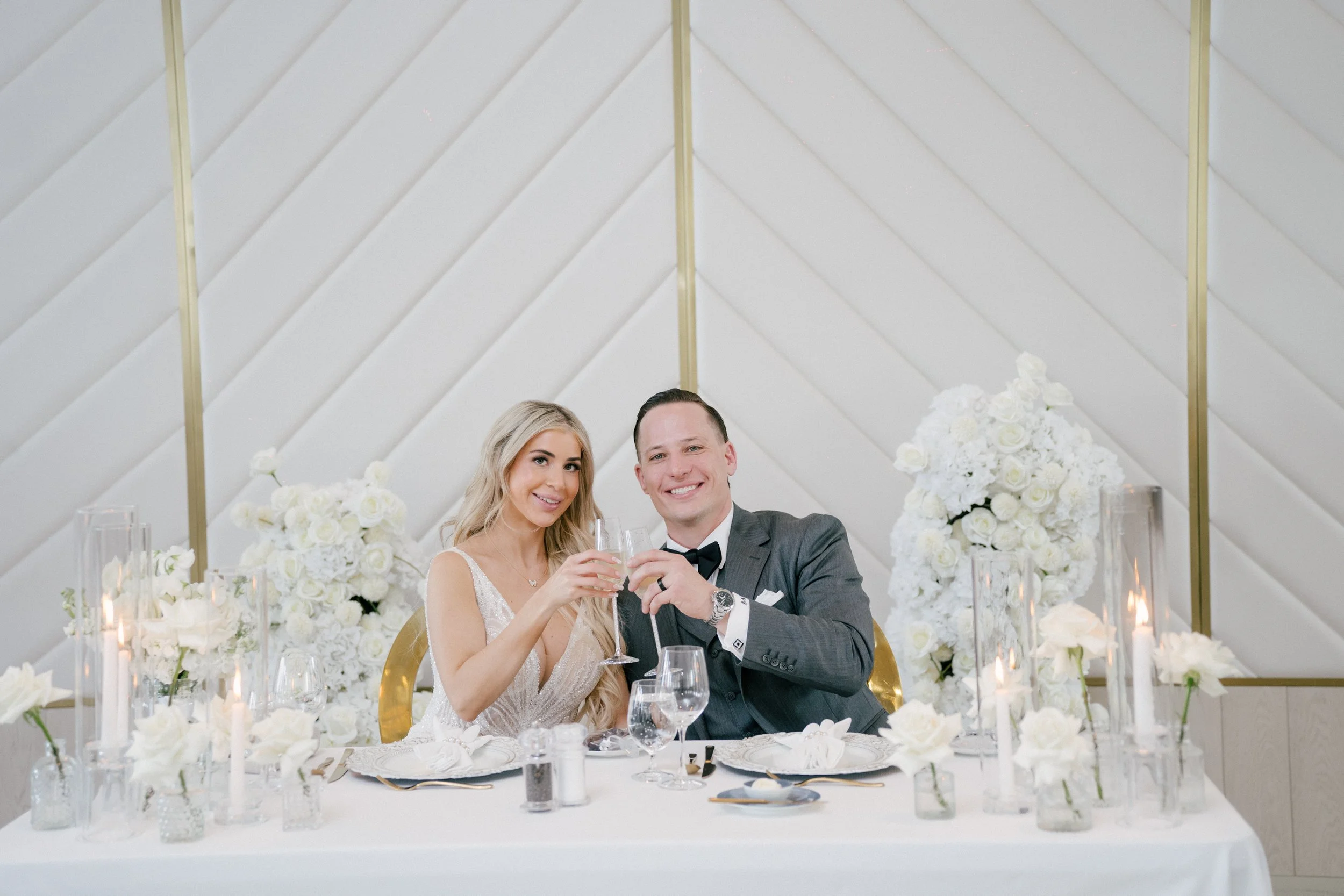 A bride and groom celebrating at their wedding reception, sitting at a decorated table with white floral arrangements, candles, and glassware, raising glasses for a toast.