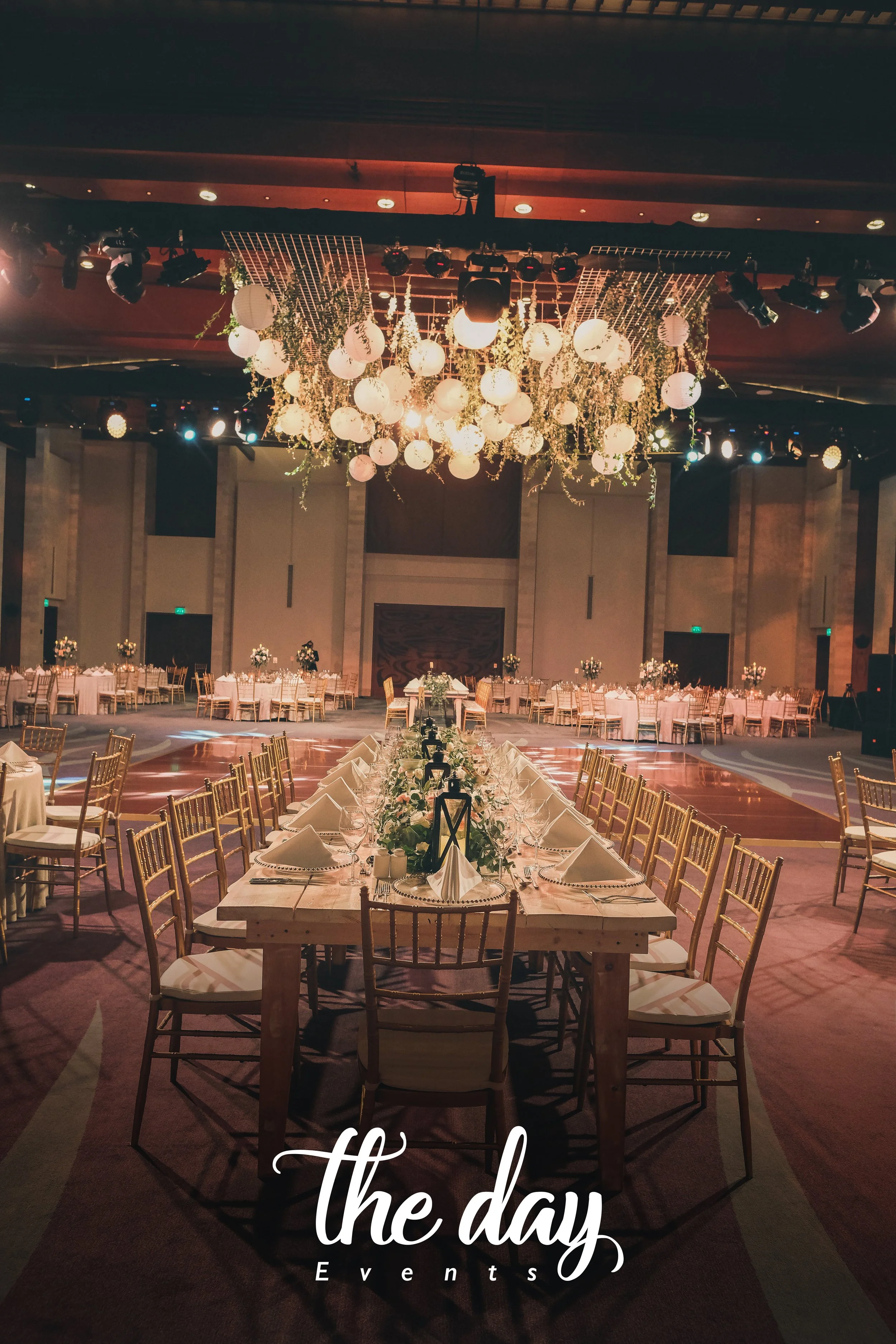 Elegant banquet hall decorated with floral arrangements, tables set with white napkins and tableware, and a large central chandelier with hanging lanterns and greenery.