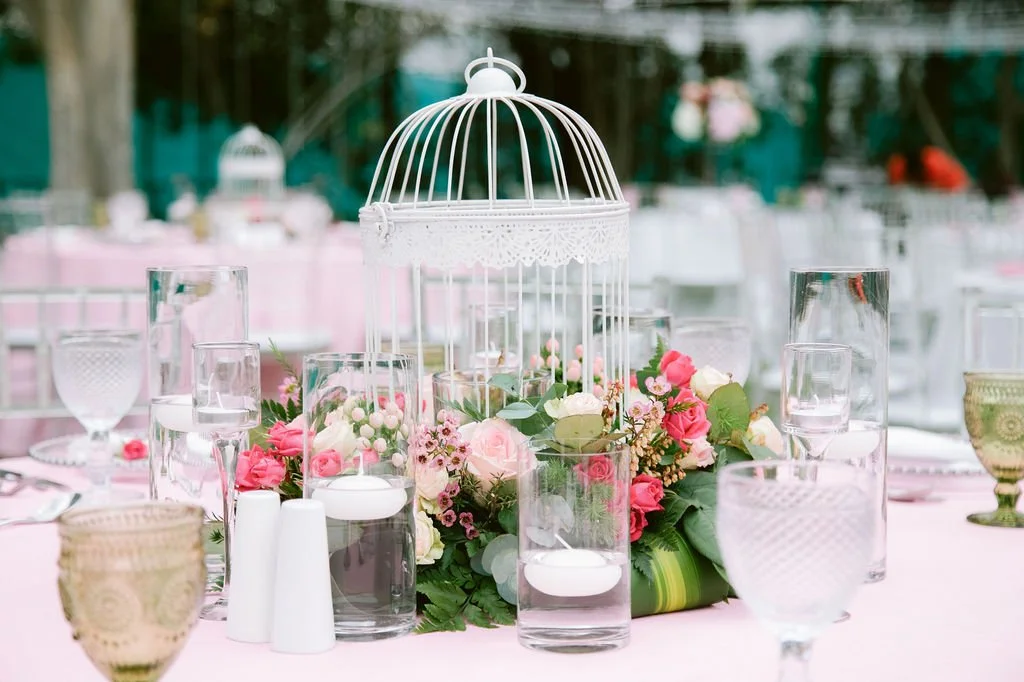 Elegant outdoor table centerpiece with pink and white flowers, glass candle holders, and a white birdcage decoration.
