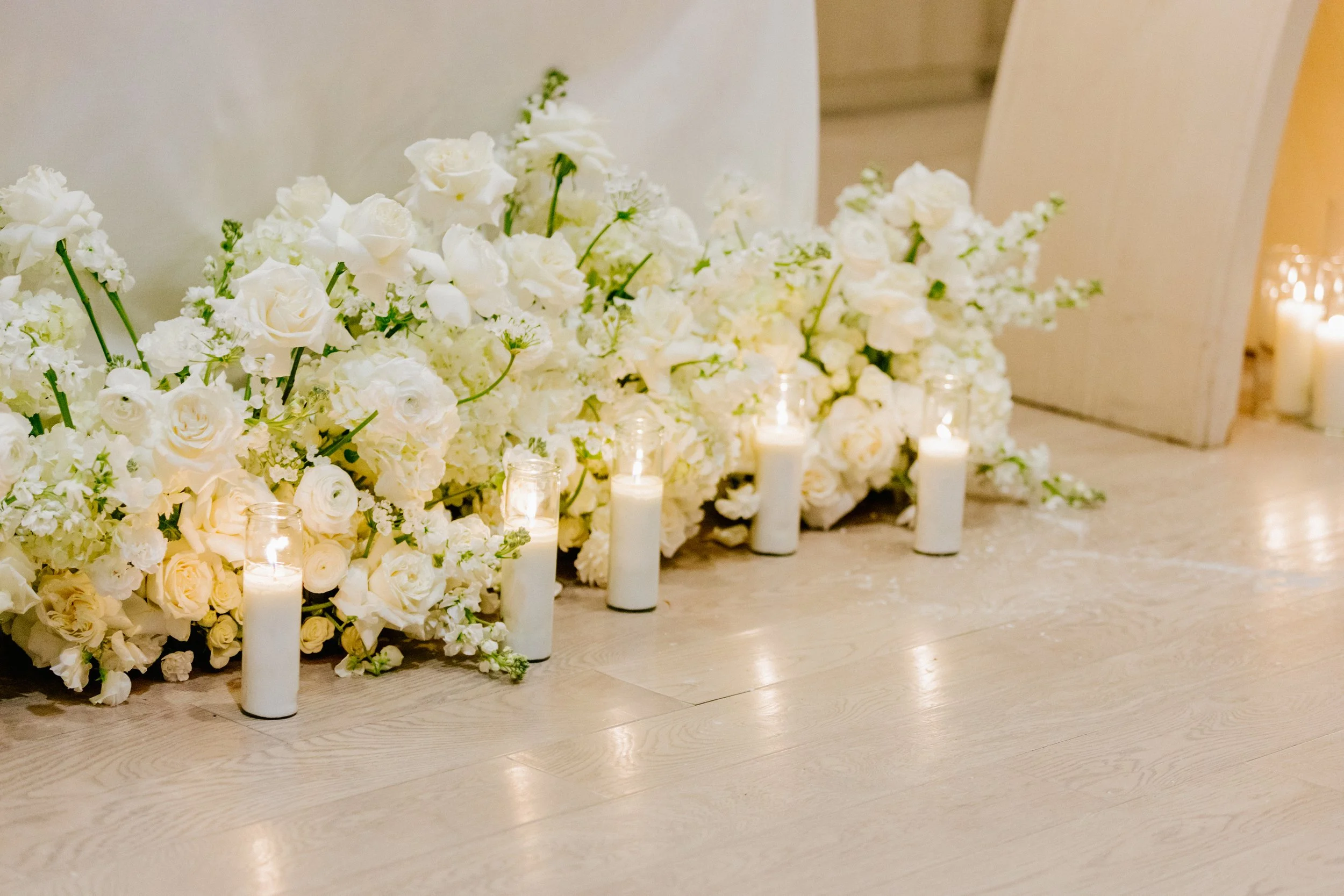 White floral arrangement with roses and lilies on a wooden floor, accented by several white candles in glass holders, in an indoor setting.