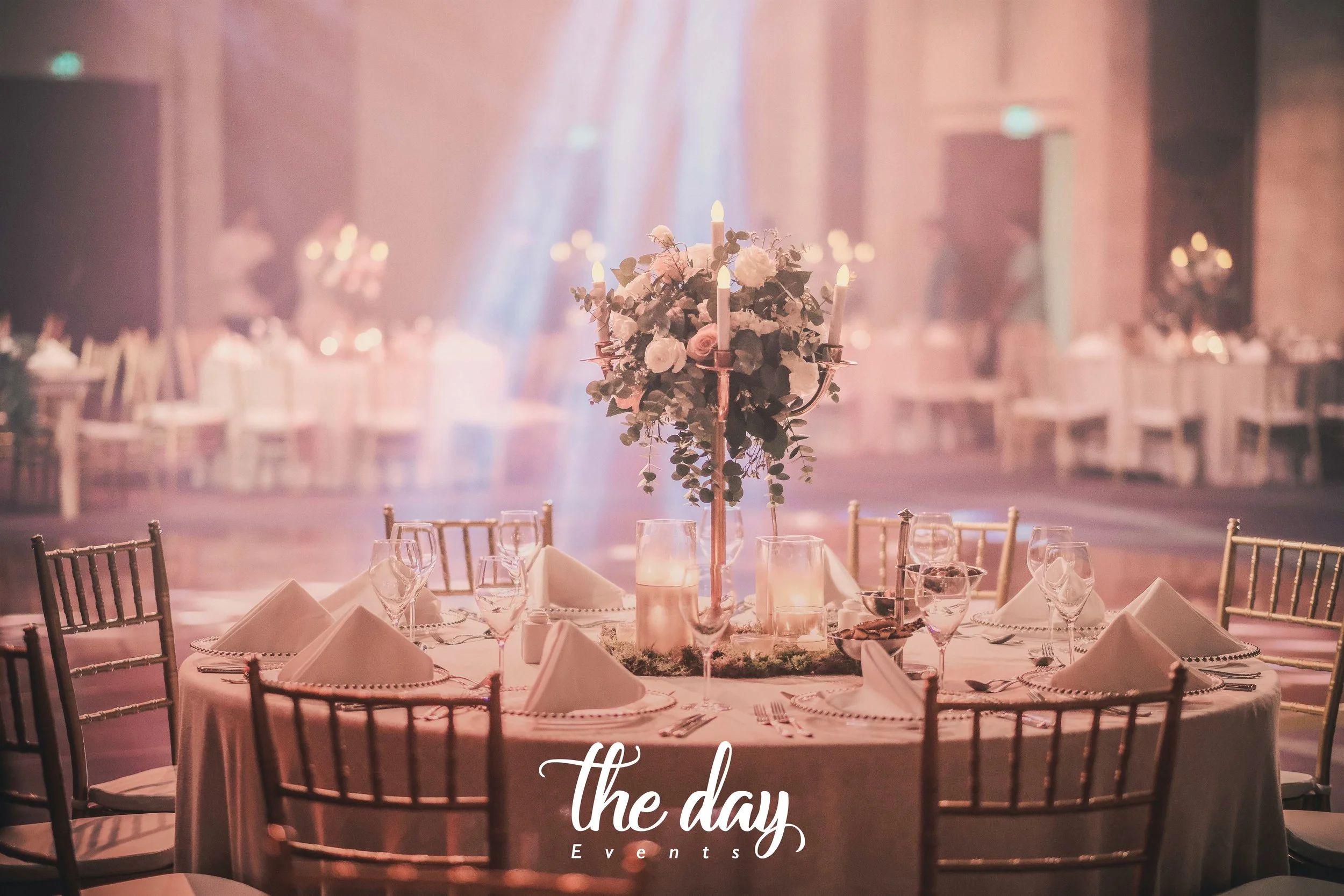 Elegant banquet table setup with a floral centerpiece, candles, wine glasses, and white napkins in a softly lit event hall.