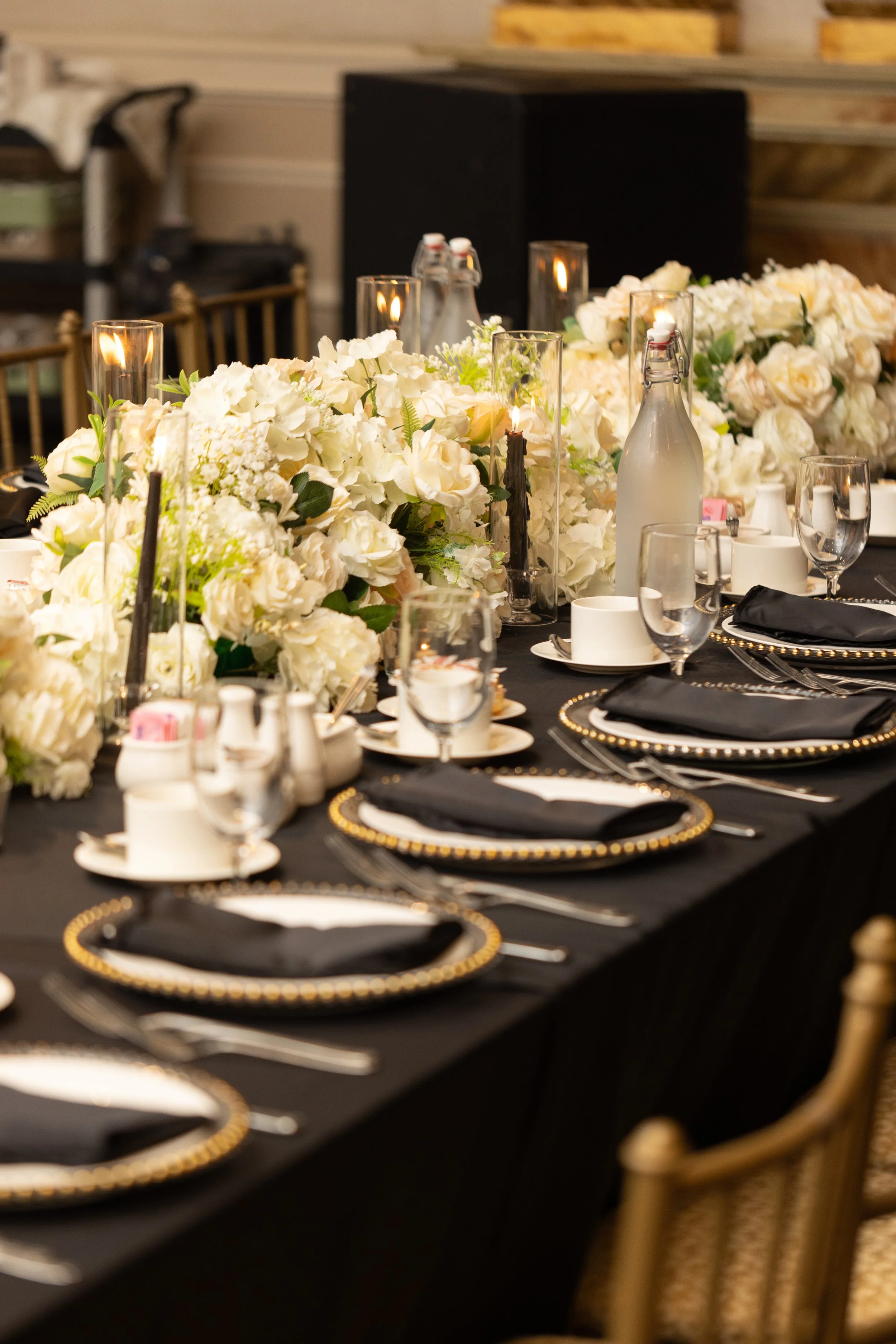 Elegant dining table set for a formal event with black and white detail including black napkins, white plates with black and gold rims, glassware, and a floral centerpiece of white roses and hydrangeas, accented with candles in glass holders.
