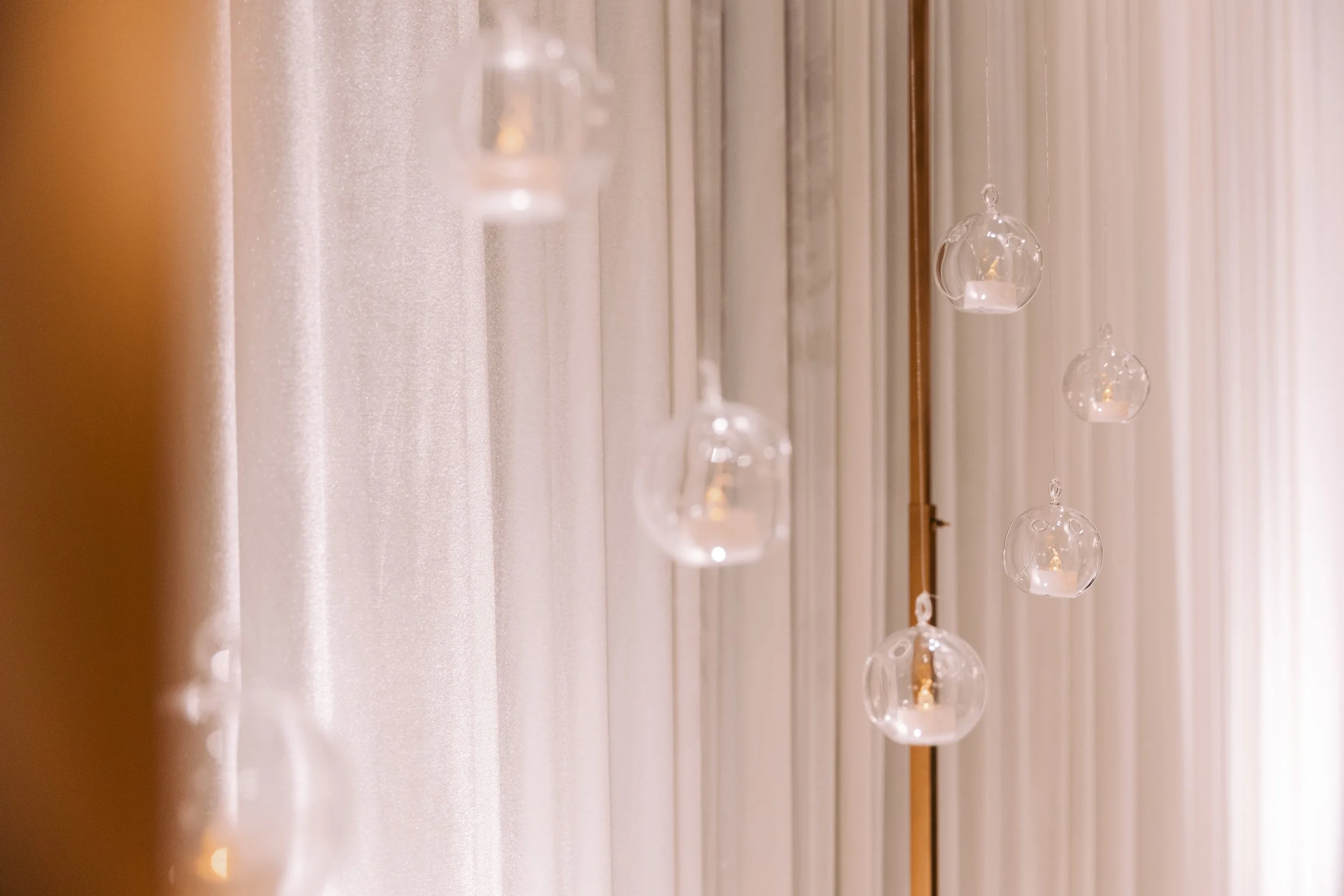Hanging glass globes with small candles inside, attached to a vertical string or wire, with vertical light-colored curtains in the background.