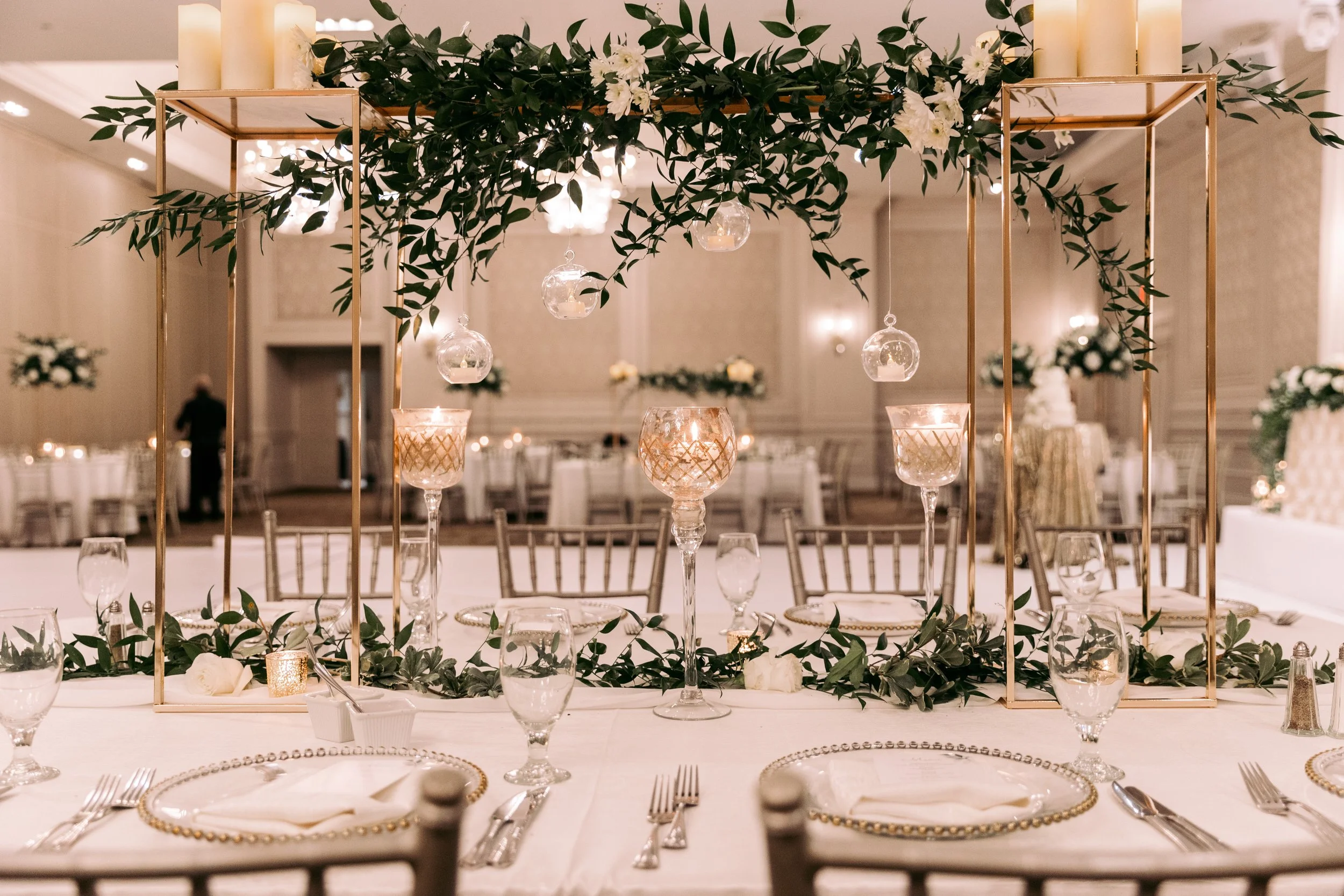 Elegant banquet table decorated with greenery, candles, and glass ornaments, featuring gold accents and place settings, set in a luxurious ballroom.