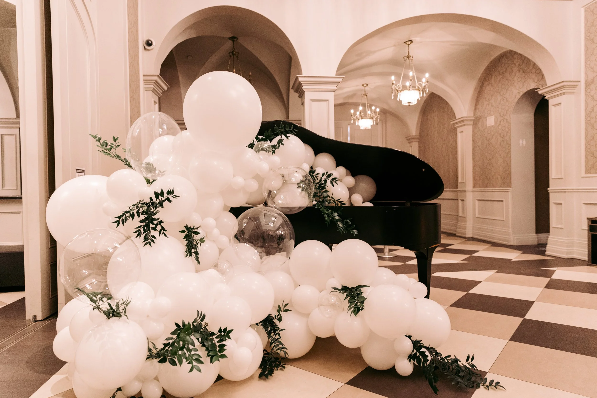 A grand piano decorated with white balloons and greenery in an elegant, well-lit interior with chandeliers and arched doorways.