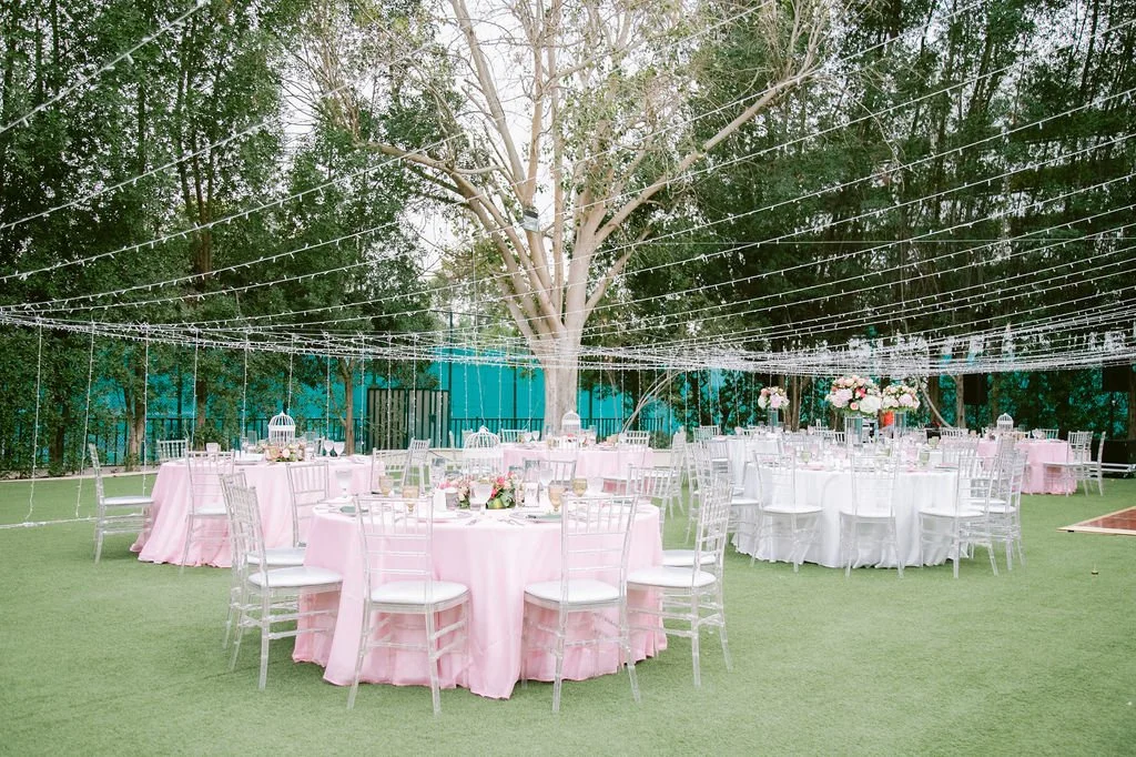 Outdoor event setup with round tables draped in pink and white tablecloths, decorated with floral centerpieces and surrounded by white chairs, under hanging string lights and a large central tree.