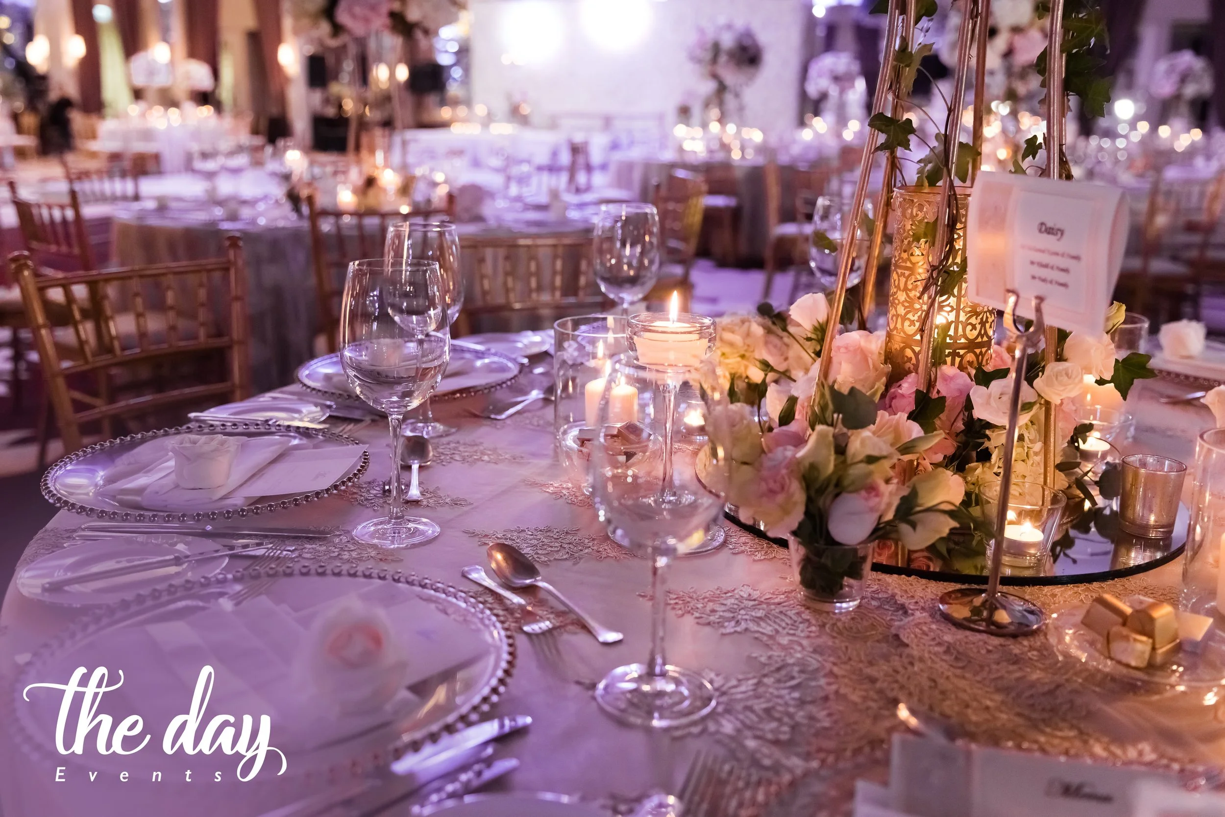 Elegant wedding reception table decor with pink and white flowers, lit candles, wine glasses, and ornate tableware in a softly lit venue.