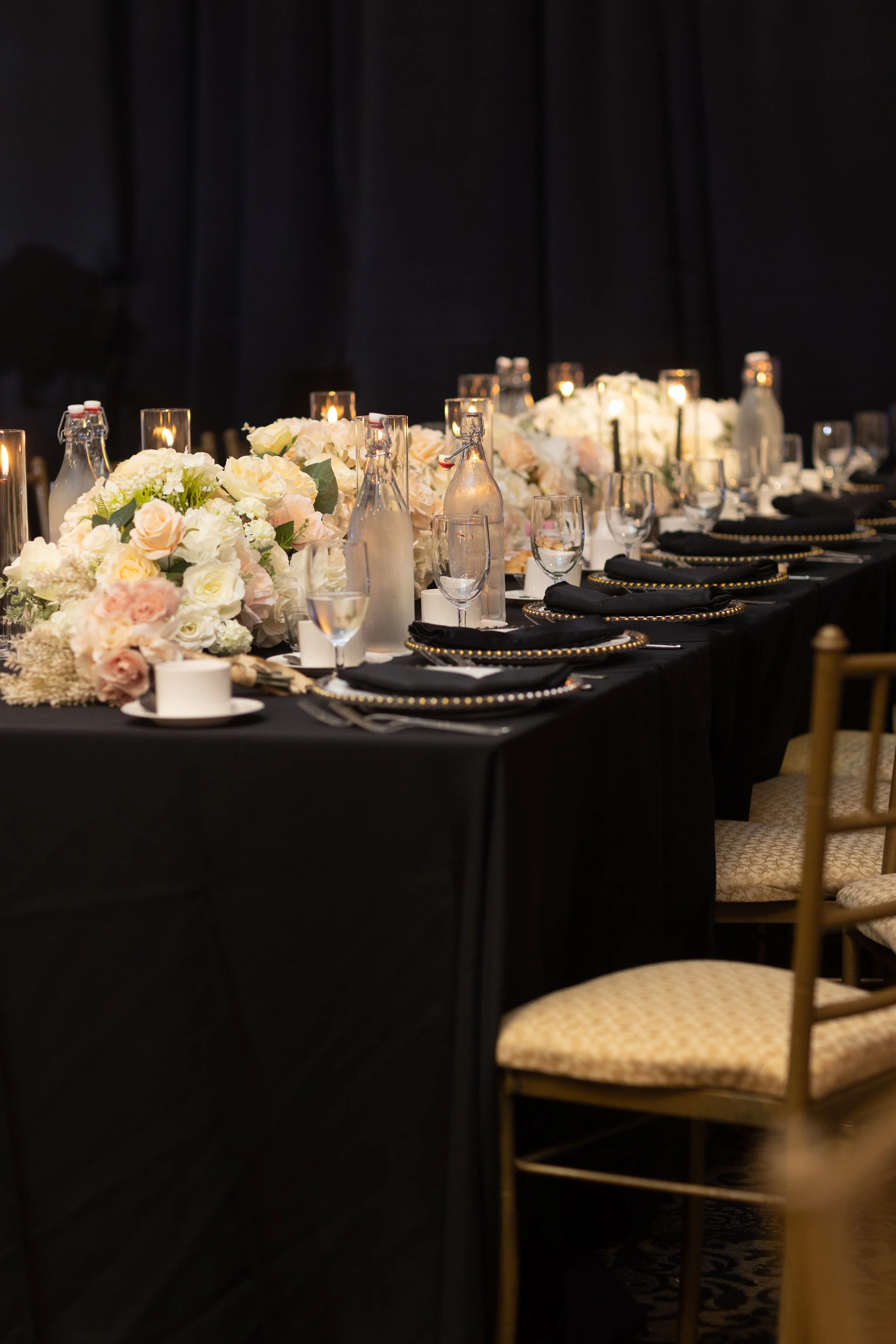 Elegant banquet table decorated with black tablecloth, floral arrangements, candles, glassware, and bottles, set against a dark backdrop.