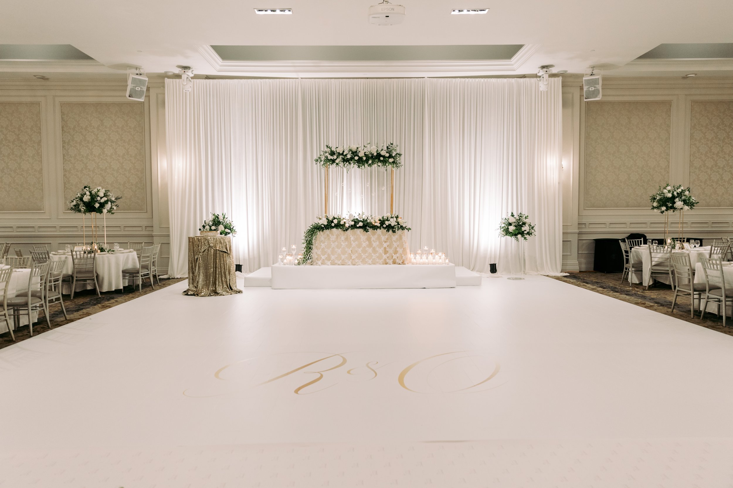 Elegant wedding reception hall with white drapery backdrop, floral arrangements, candles, and monogrammed dance floor.
