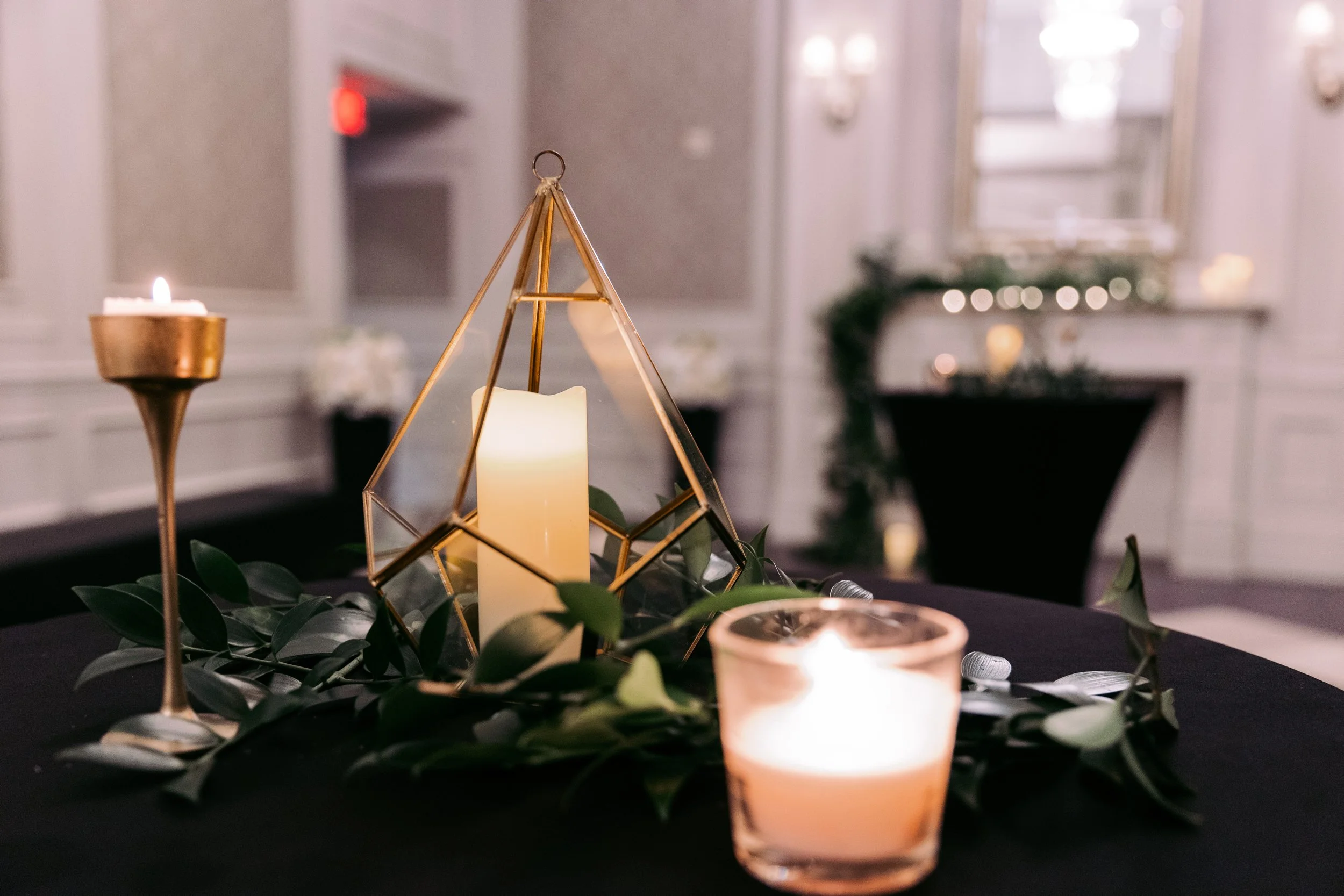 Central candle holder with a pillar candle inside a geometric glass and metal frame, surrounded by greenery and smaller candles, on a black table in an elegant, well-lit room.