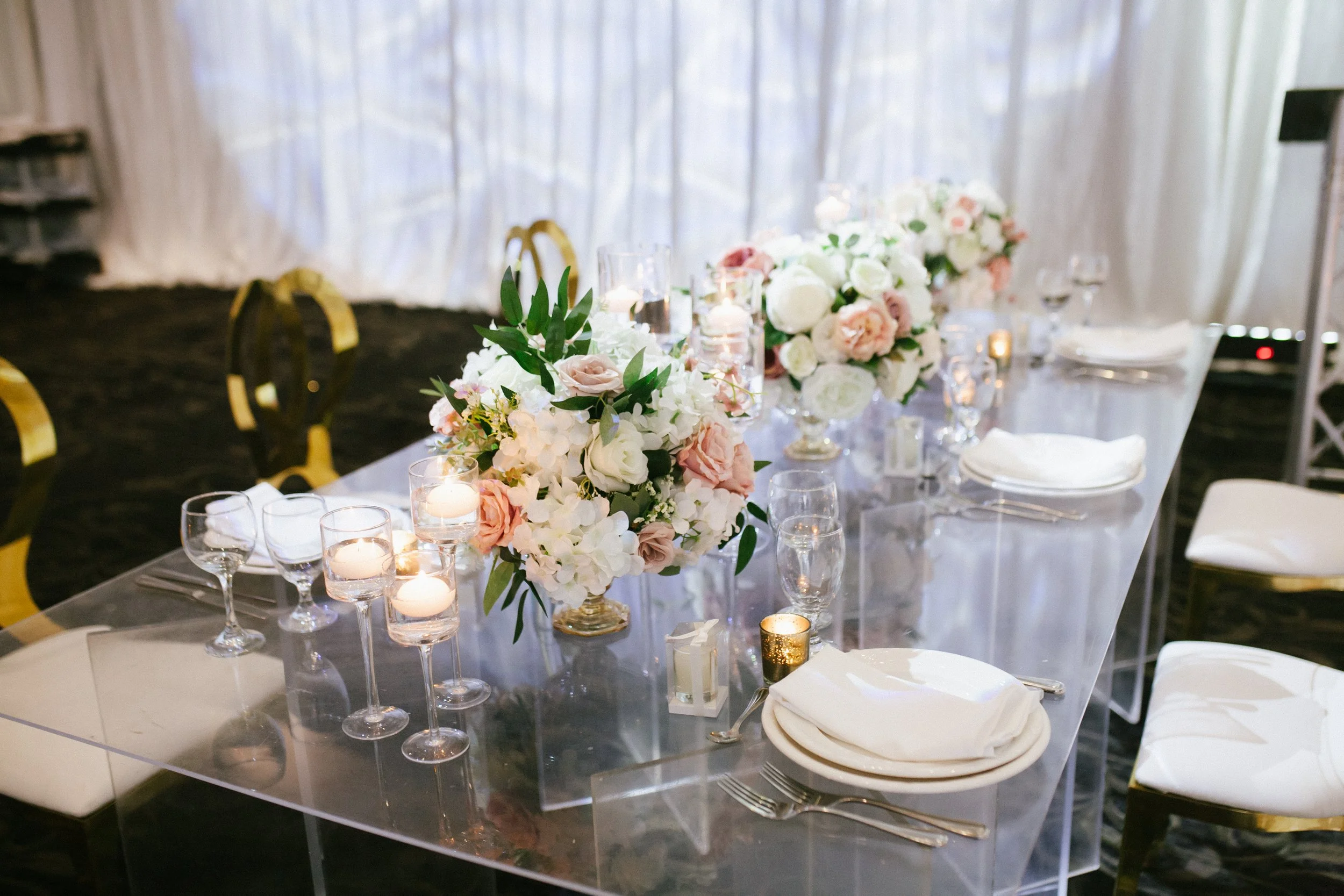 Elegant wedding reception table with white and blush floral centerpieces, lit candles in glass holders, white plates, napkins, and silverware, clear chairs with gold accents, and a white backdrop.