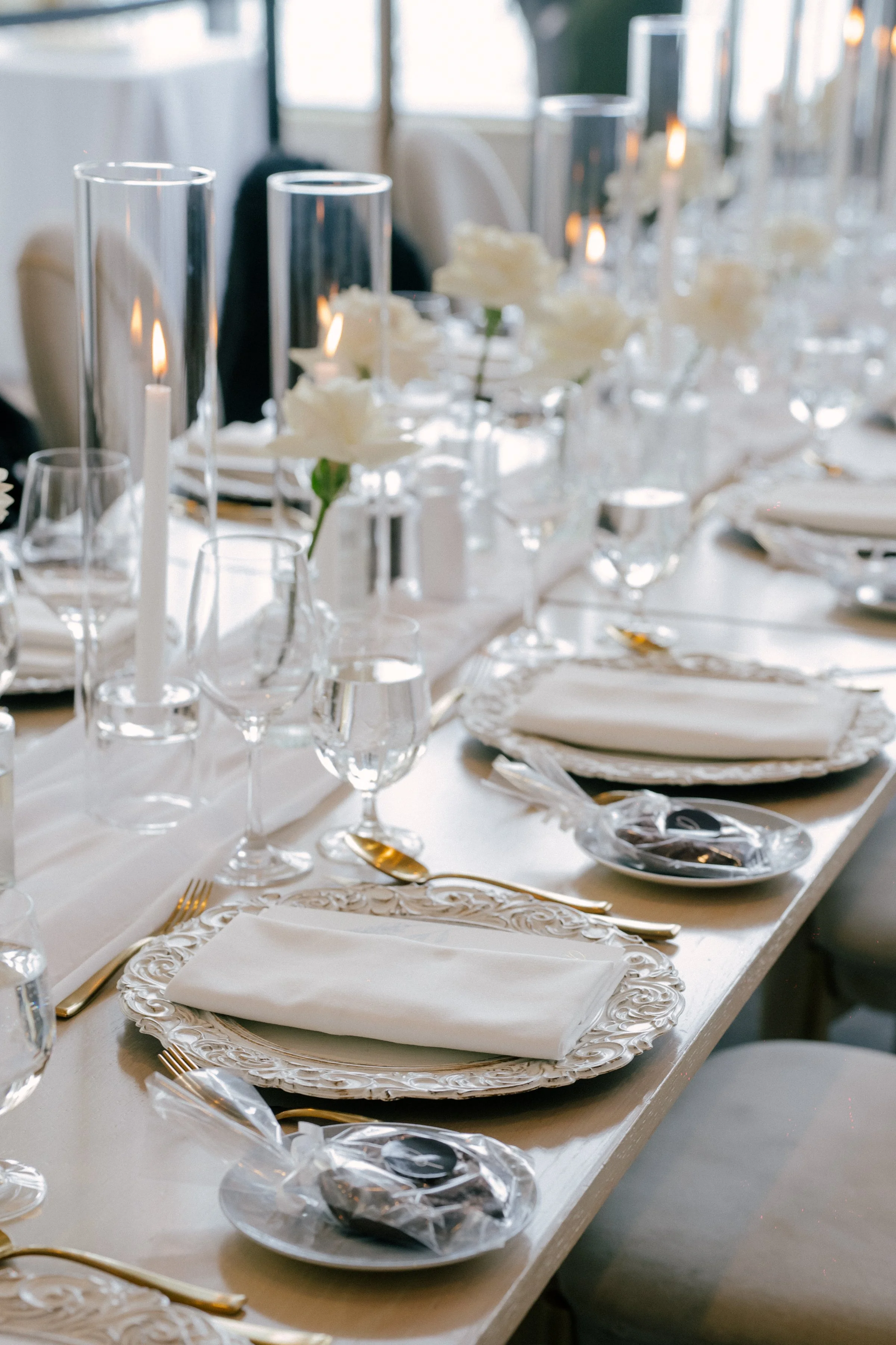 Elegant banquet table set with white plates, linen napkins, gold utensils, and glassware, decorated with white flowers and tall glass candle holders.