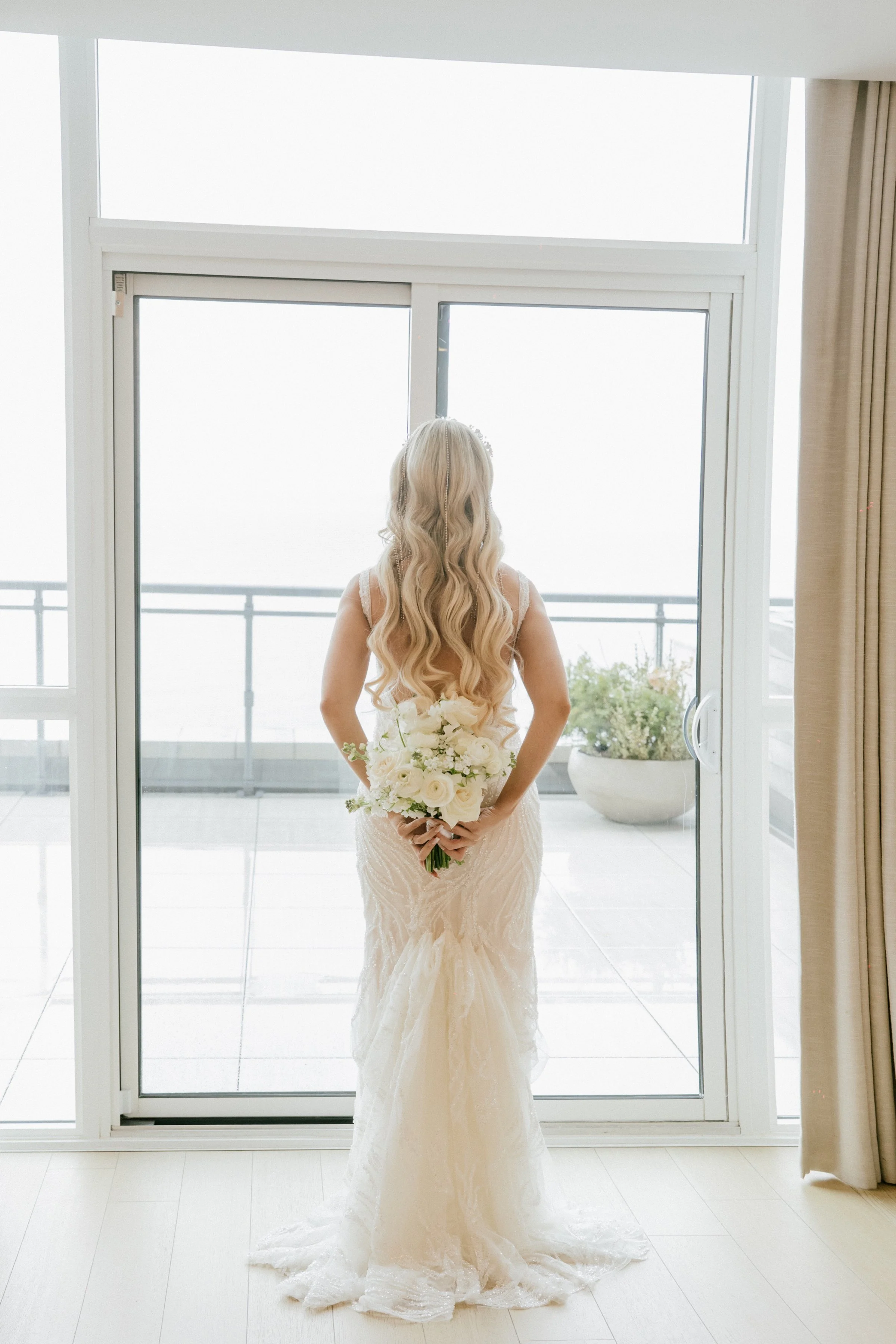 Bride with long blonde hair in a white wedding dress holding a bouquet standing in front of glass sliding doors leading to a balcony.