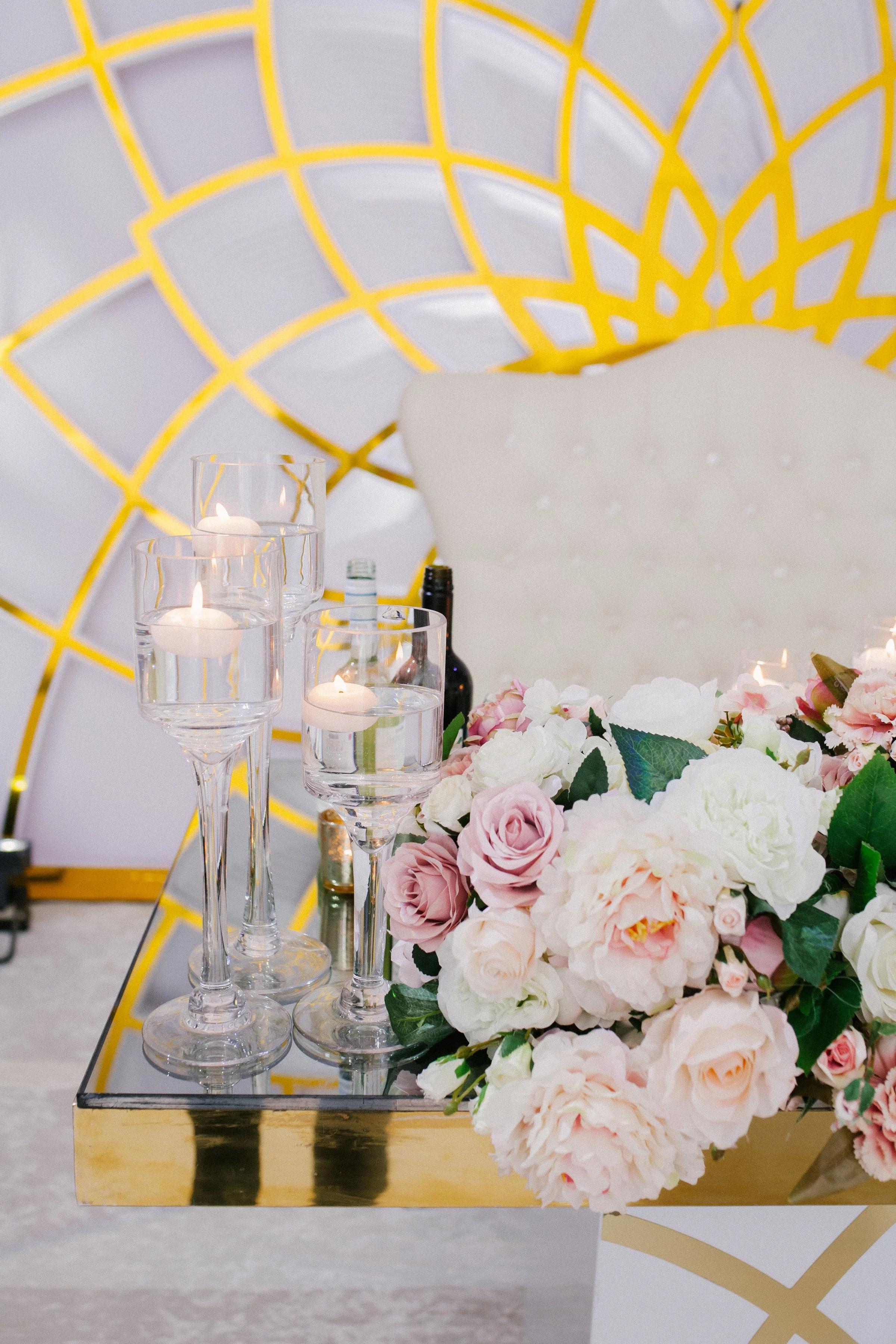 A table decorated with a bouquet of pink and white roses, three floating candles in glass holders, and bottles of alcohol, set against a gold and white geometric backdrop.