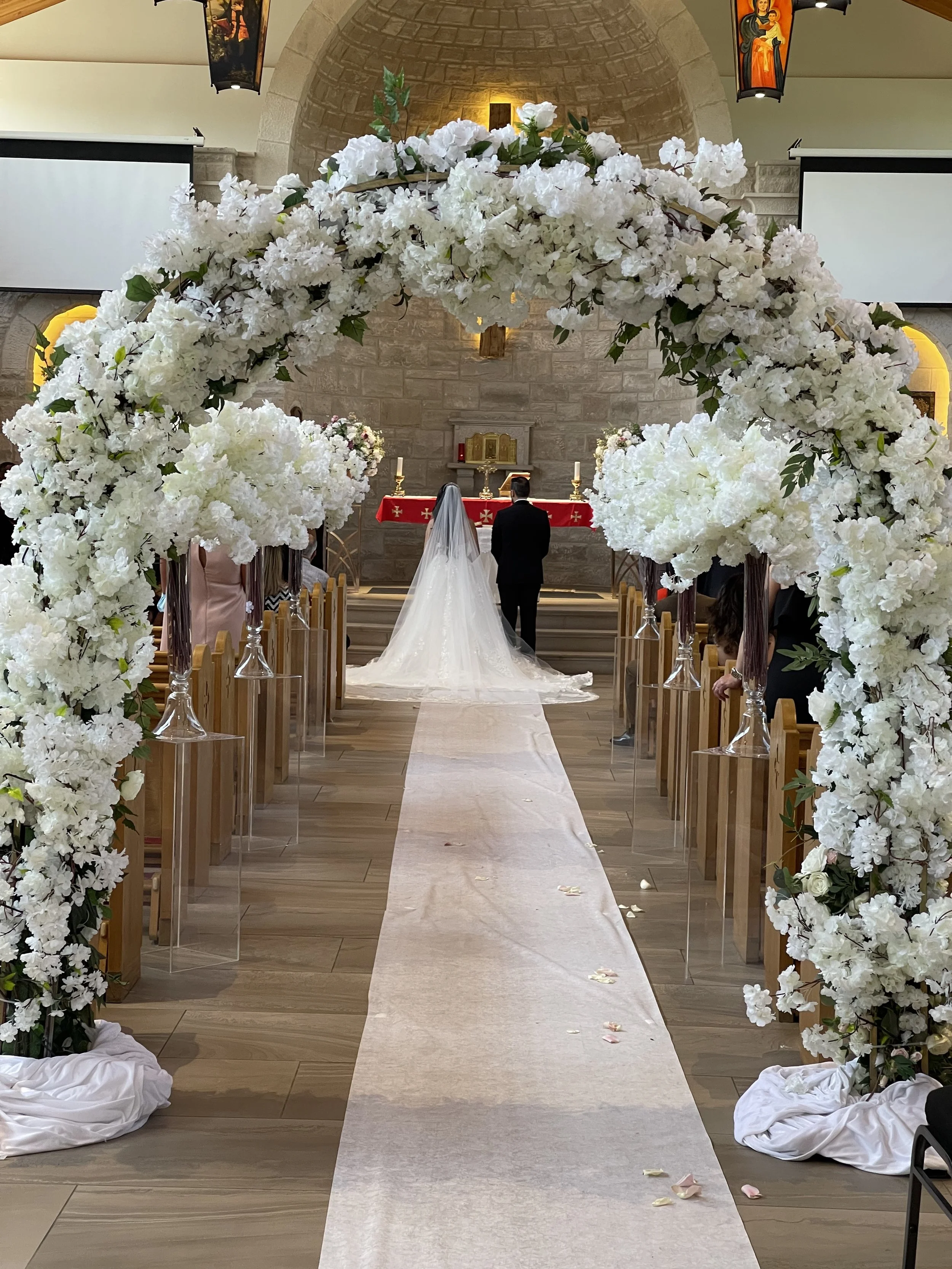 Wedding ceremony inside a church with a floral archway framing the aisle, a bride and groom standing at the altar, and guests seated on either side.