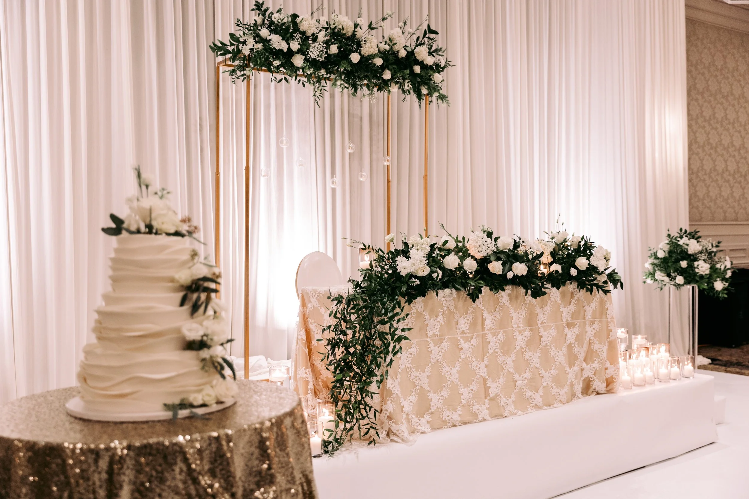 Wedding reception table decorated with white roses, greenery, and candles; a tiered cake decorated with white flowers in the foreground.