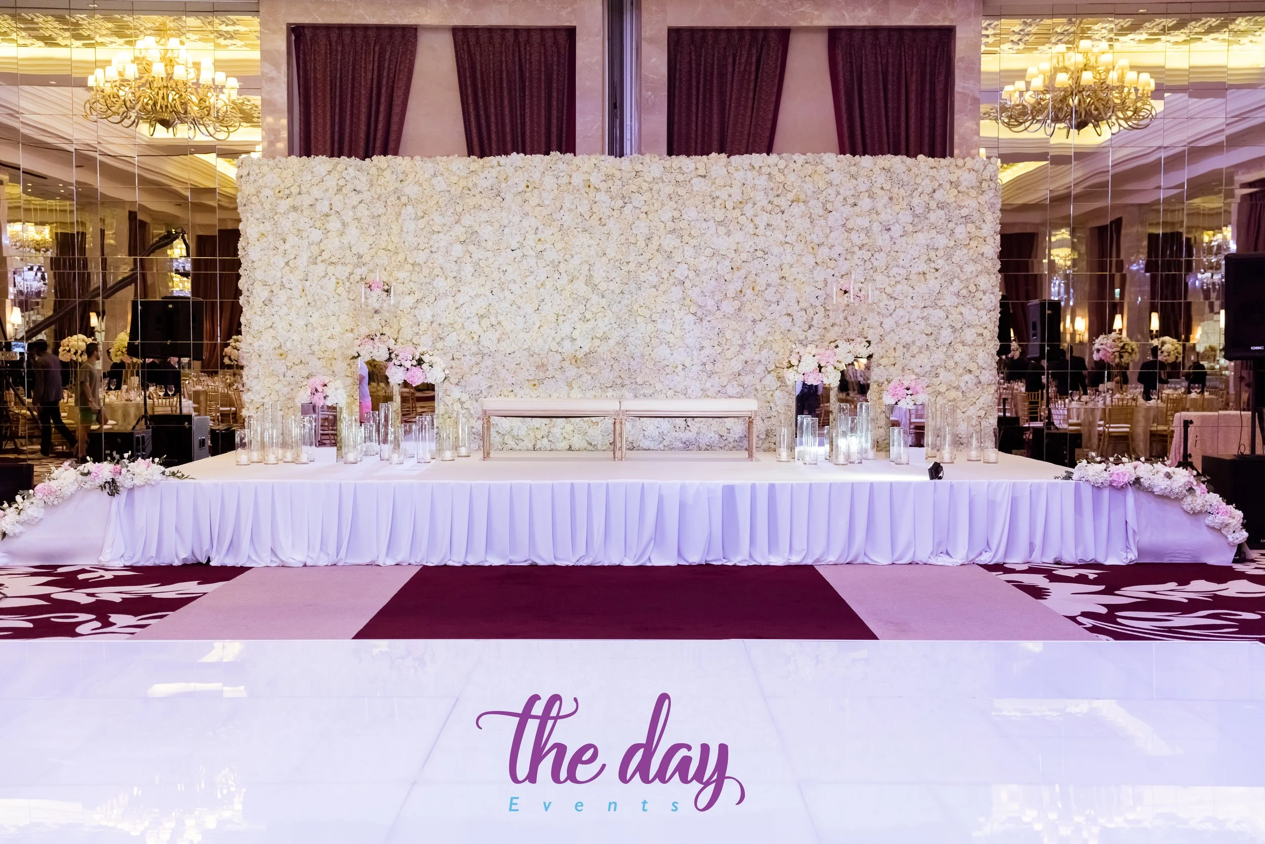 A decorated stage with a large white floral backdrop, surrounded by vases with pink and white flowers, set for a wedding or event at a banquet hall with chandeliers and mirrored walls.