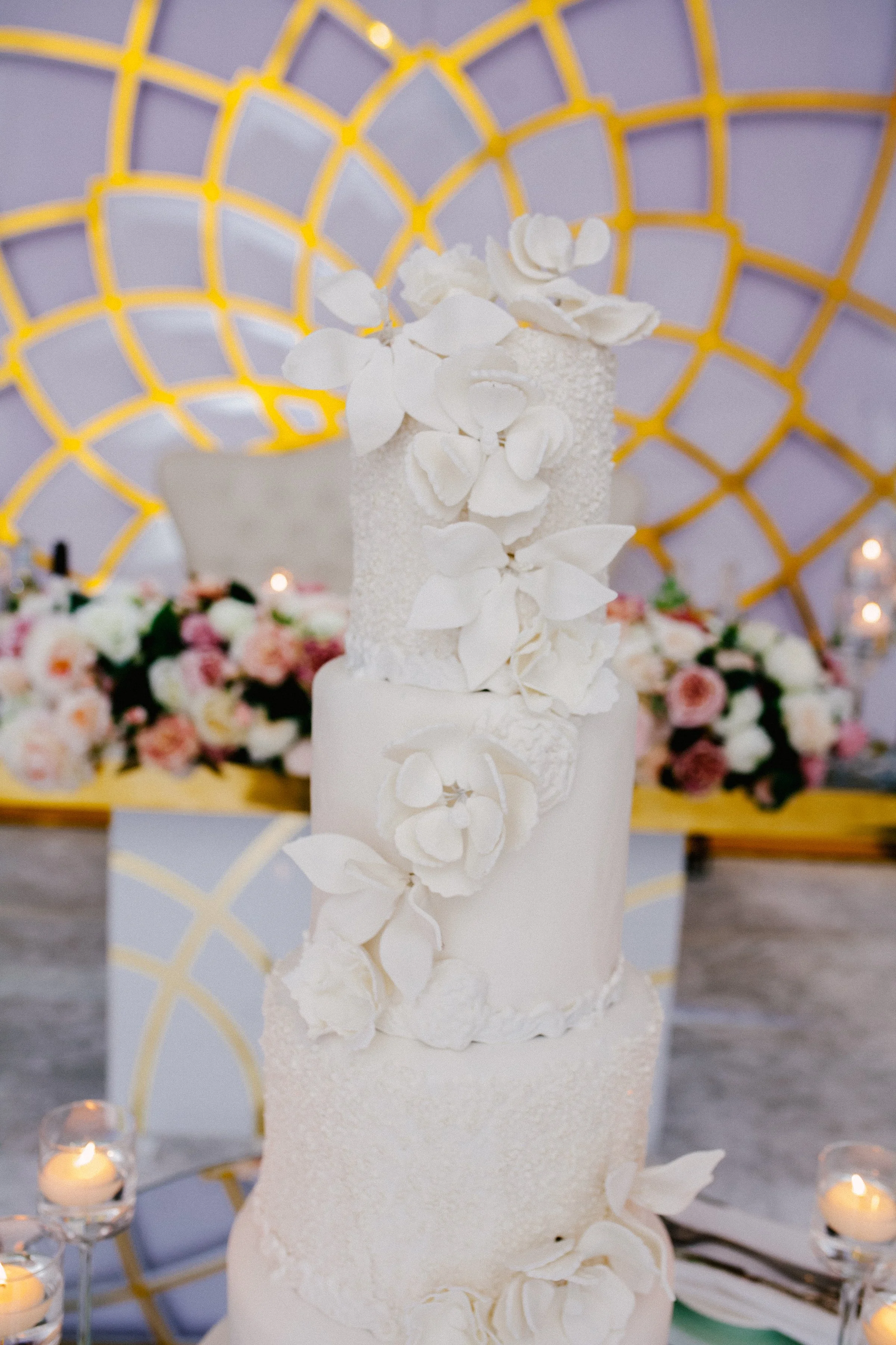 A wedding cake with three tiers, decorated with white flowers and leaves, and a floral arrangement with candles in the background.