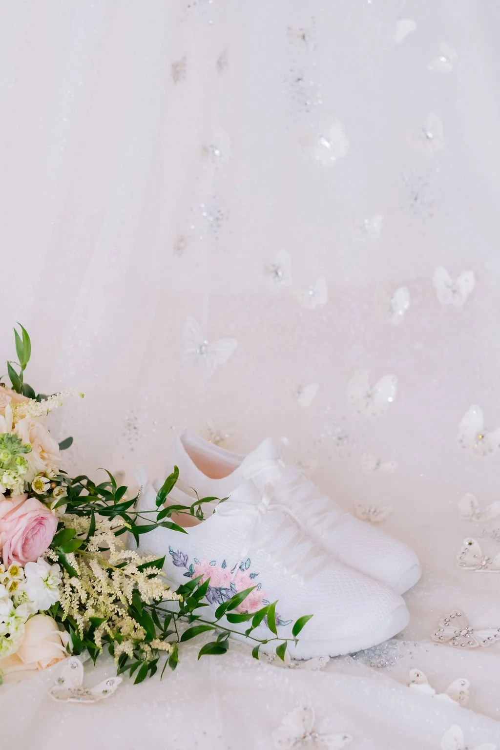 White athletic shoes with floral embroidery, a bouquet of flowers with pink roses and greenery, and a backdrop with white butterfly and heart-shaped embellishments.