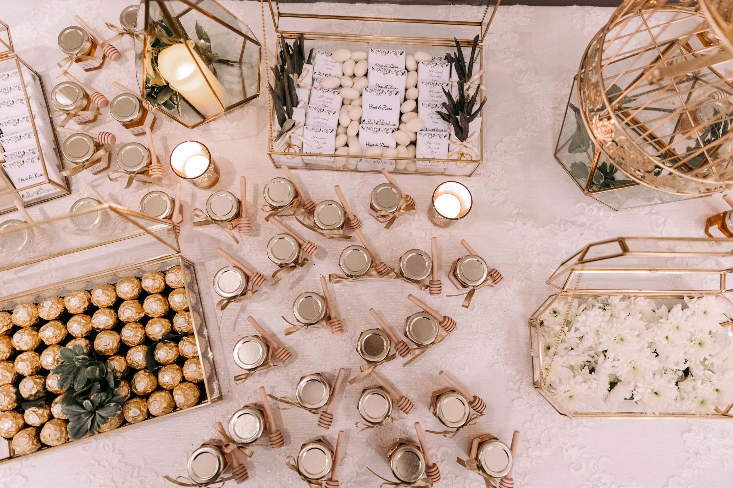 Elegant table display with white almonds, wedding favors, and small jars with gold ribbon and lids, decorated with candles and floral arrangements in gold geometric containers.