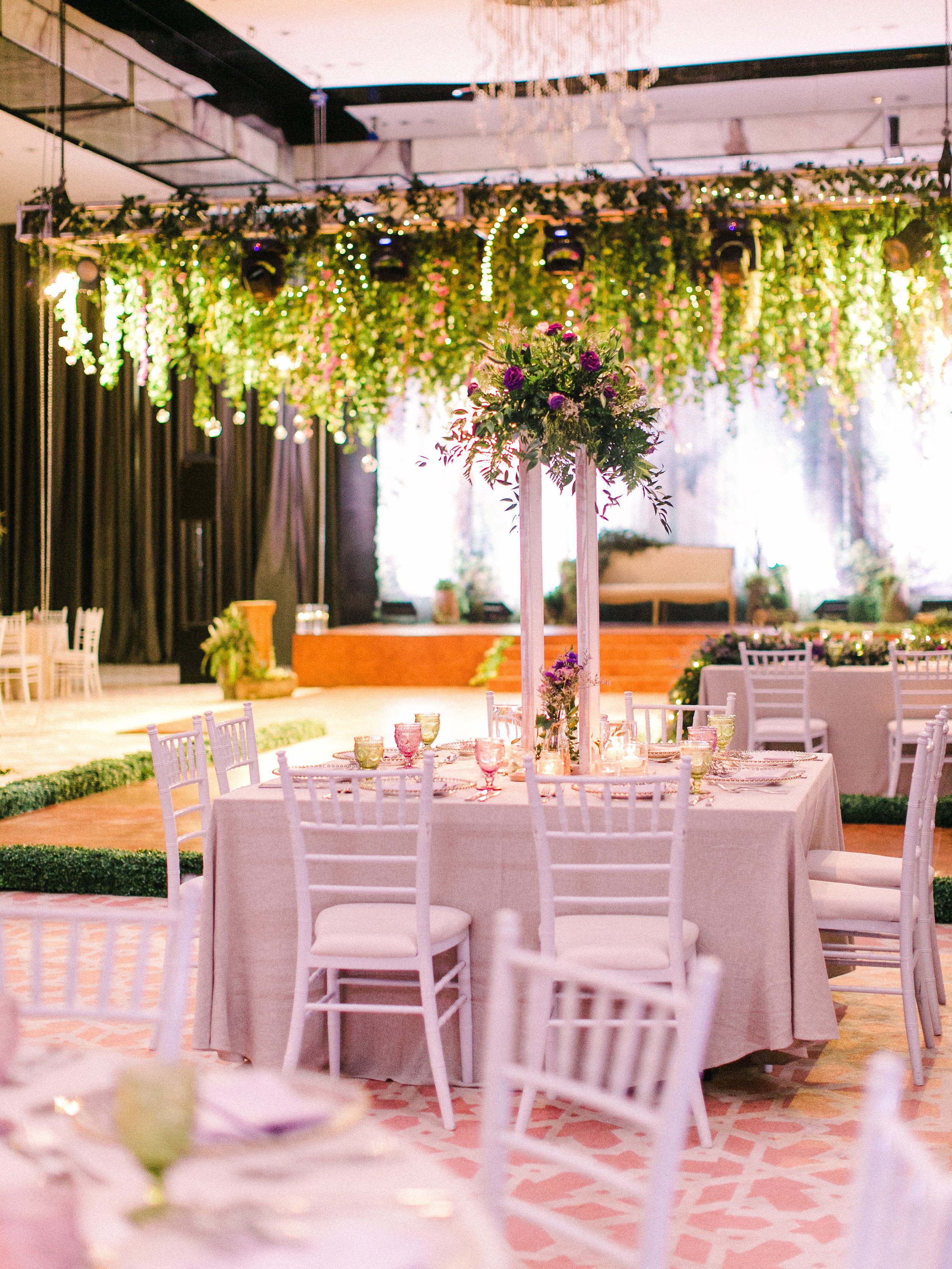 Elegant event space with round tables, white chairs, floral centerpieces, and hanging greenery with fairy lights.