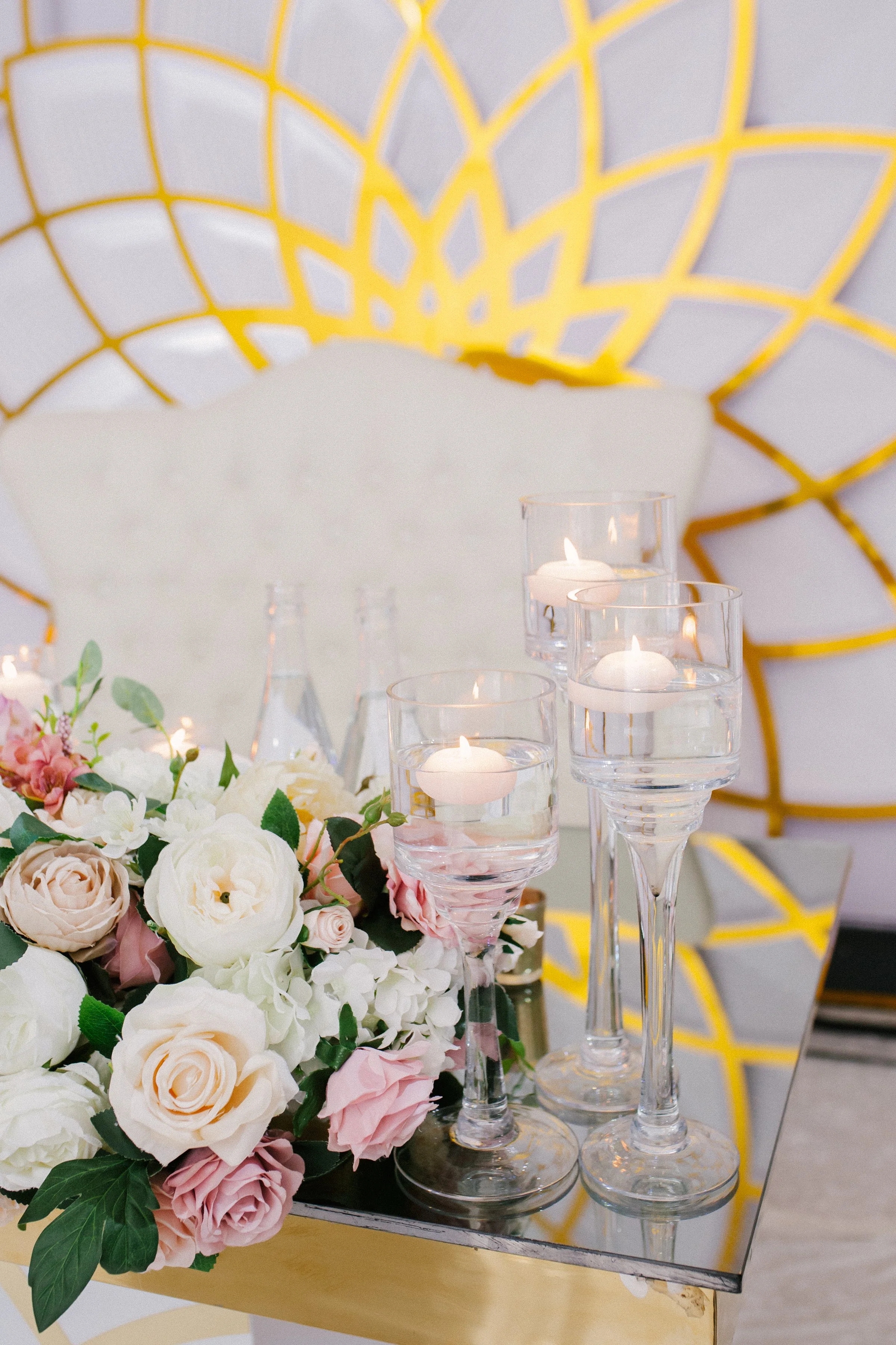 A floral centerpiece with white and pink roses and greenery, four glass candle holders with floating candles, set on a mirrored table, with a yellow decorative wall in the background.