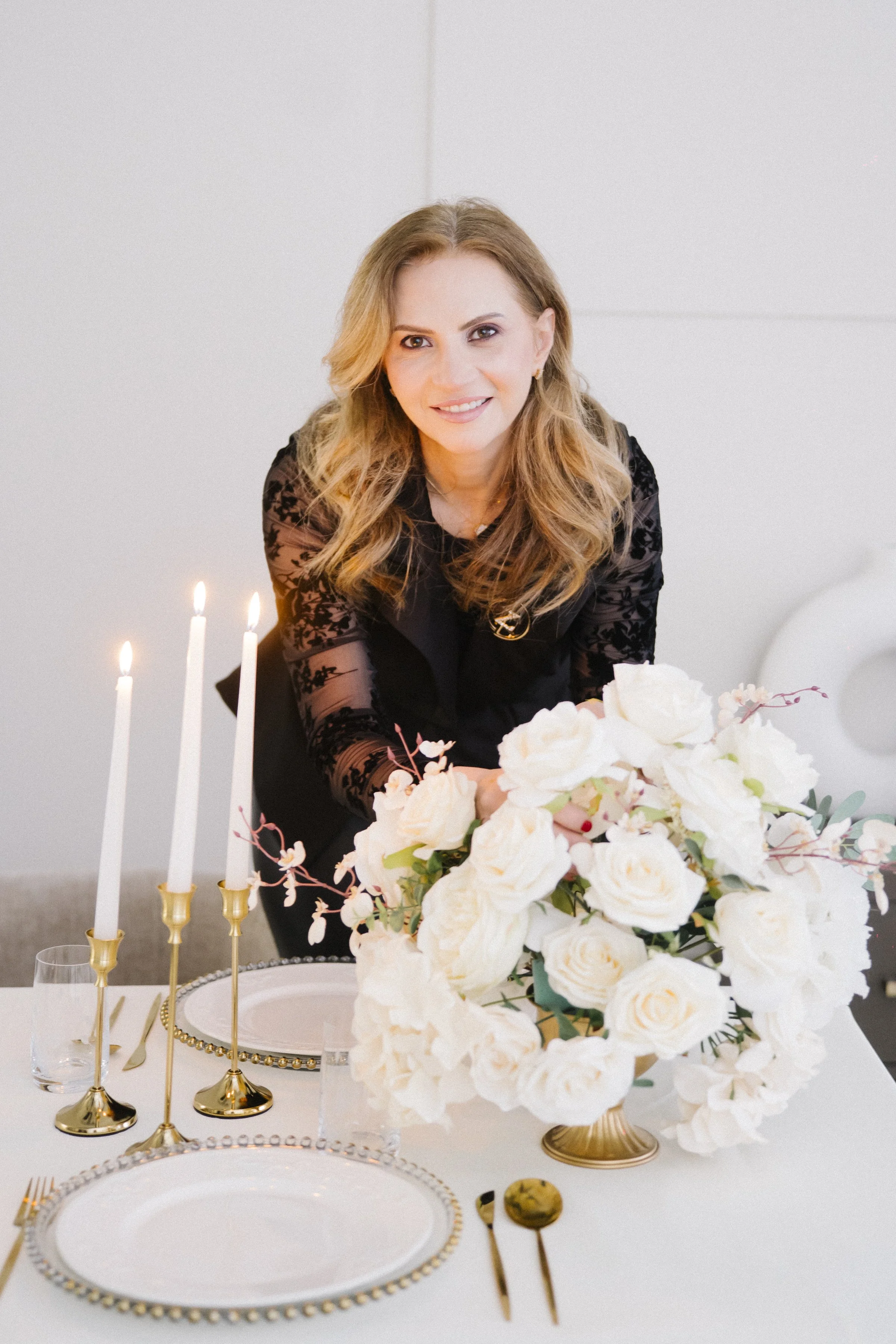 A woman with wavy blonde hair smiling at a decorated dinner table with white roses, candles, and gold-rimmed plates.